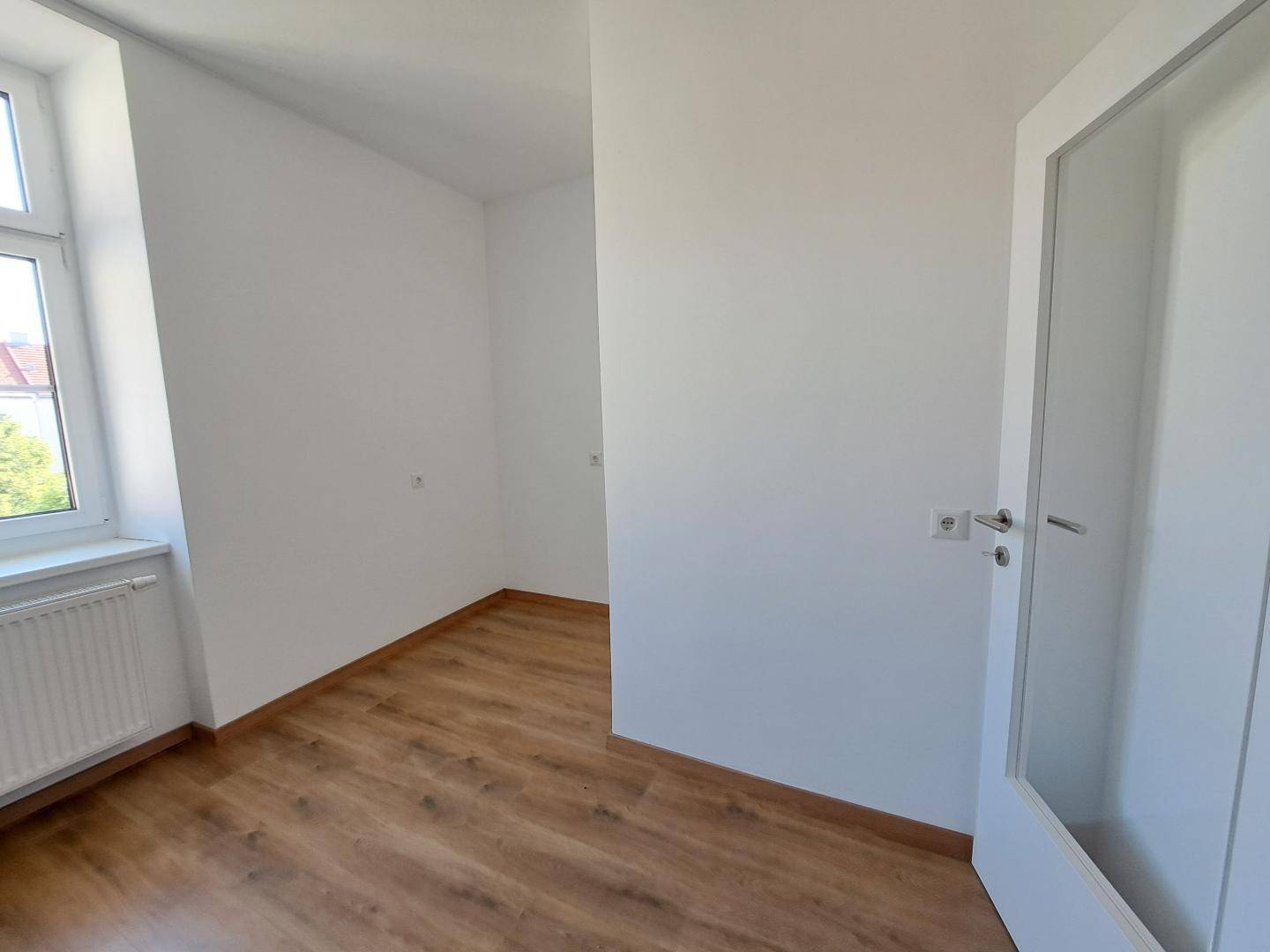 Heller, leerer Raum mit Laminatboden und einem Fenster, das Tageslicht hereinlässt.