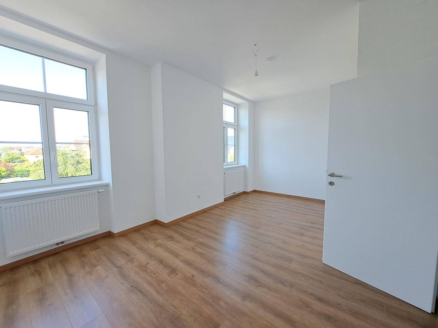 Lichtdurchfluteter Raum mit zwei großen Fenstern und Laminatboden, ideal für ein Wohnzimmer.