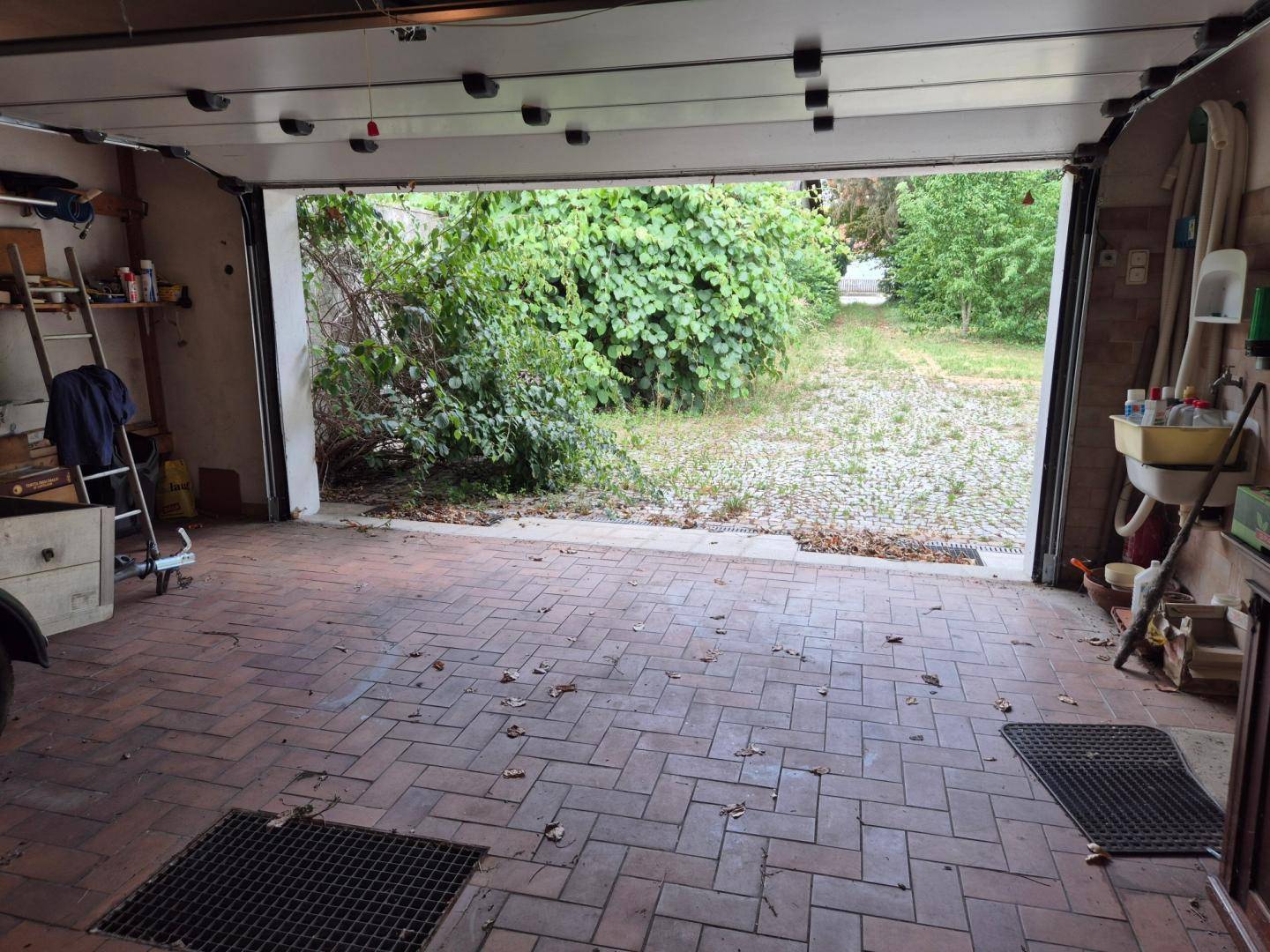 Geräumige Garage mit Ziegelboden und Blick auf den verwilderten Außenbereich.