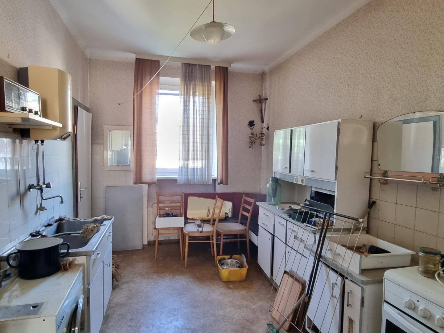 Veraltete Küche mit einfachen Schränken, Spüle und kleinem Essbereich am Fenster in originalem Zustand.