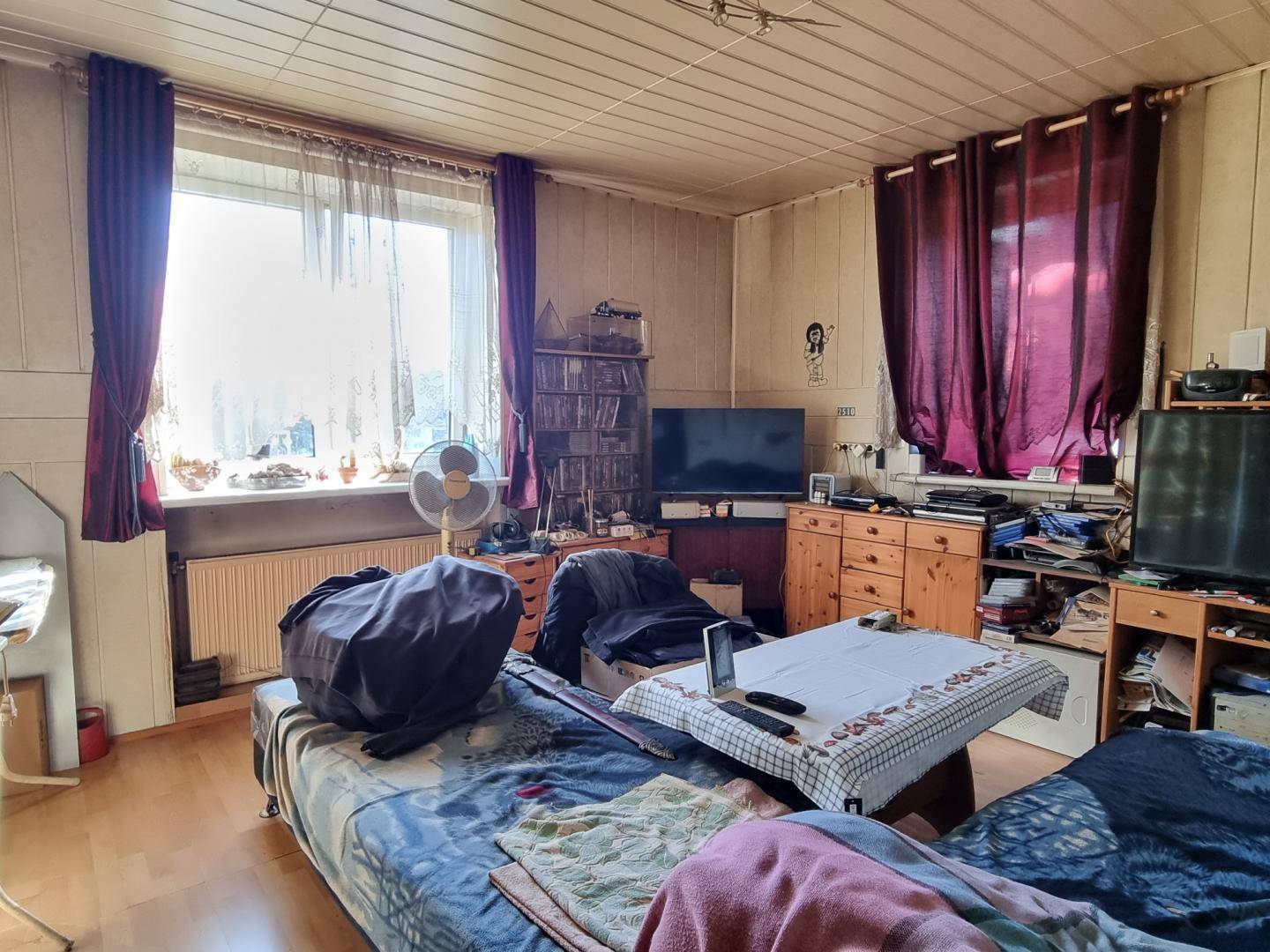 Unaufgeräumtes Schlafzimmer mit großem Fenster, Vorhängen und älteren Möbeln in originalem Zustand.