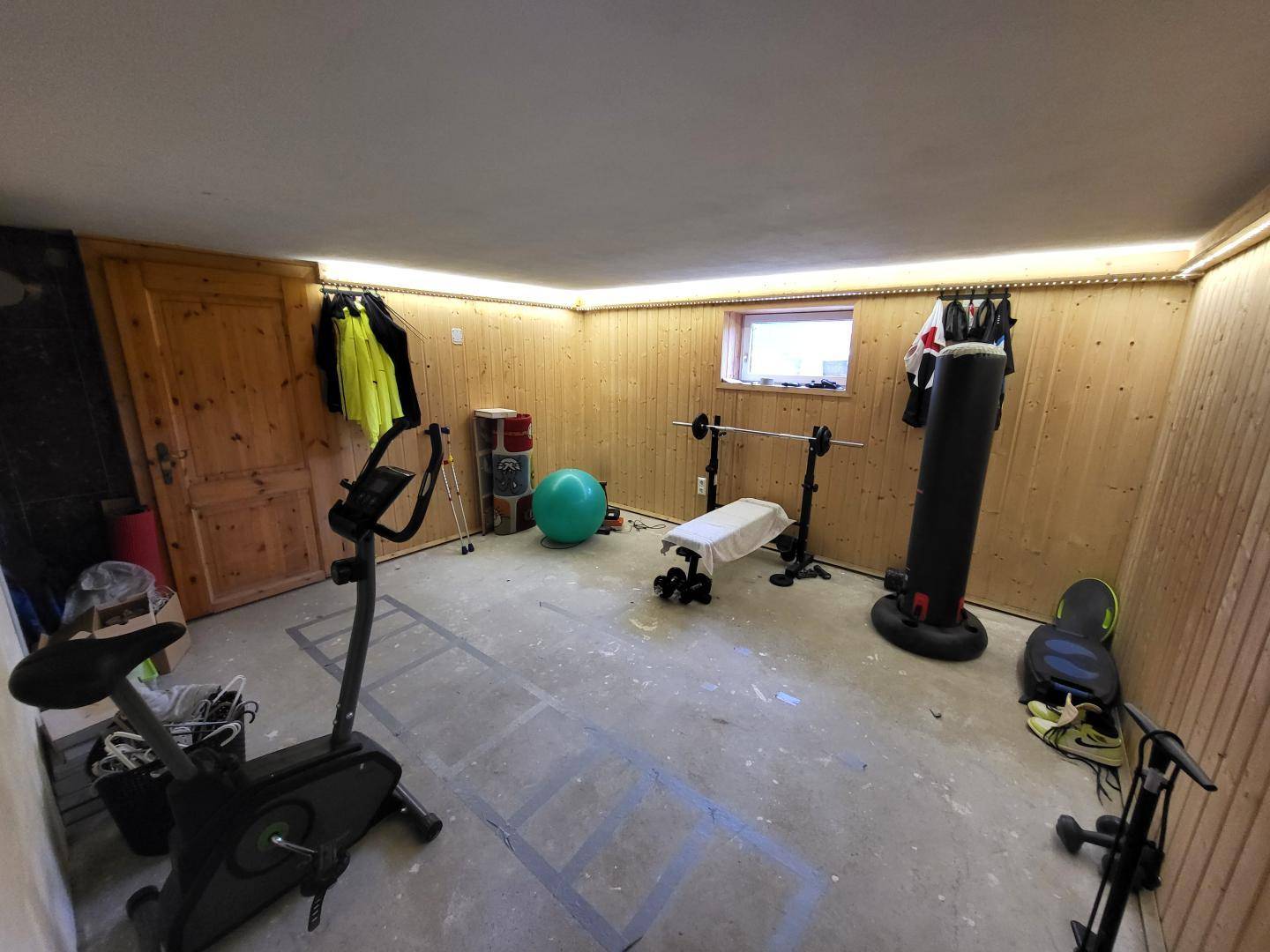 Fitnessraum im Keller mit Holzverkleidung, Sportgeräten und kleinem Fenster.