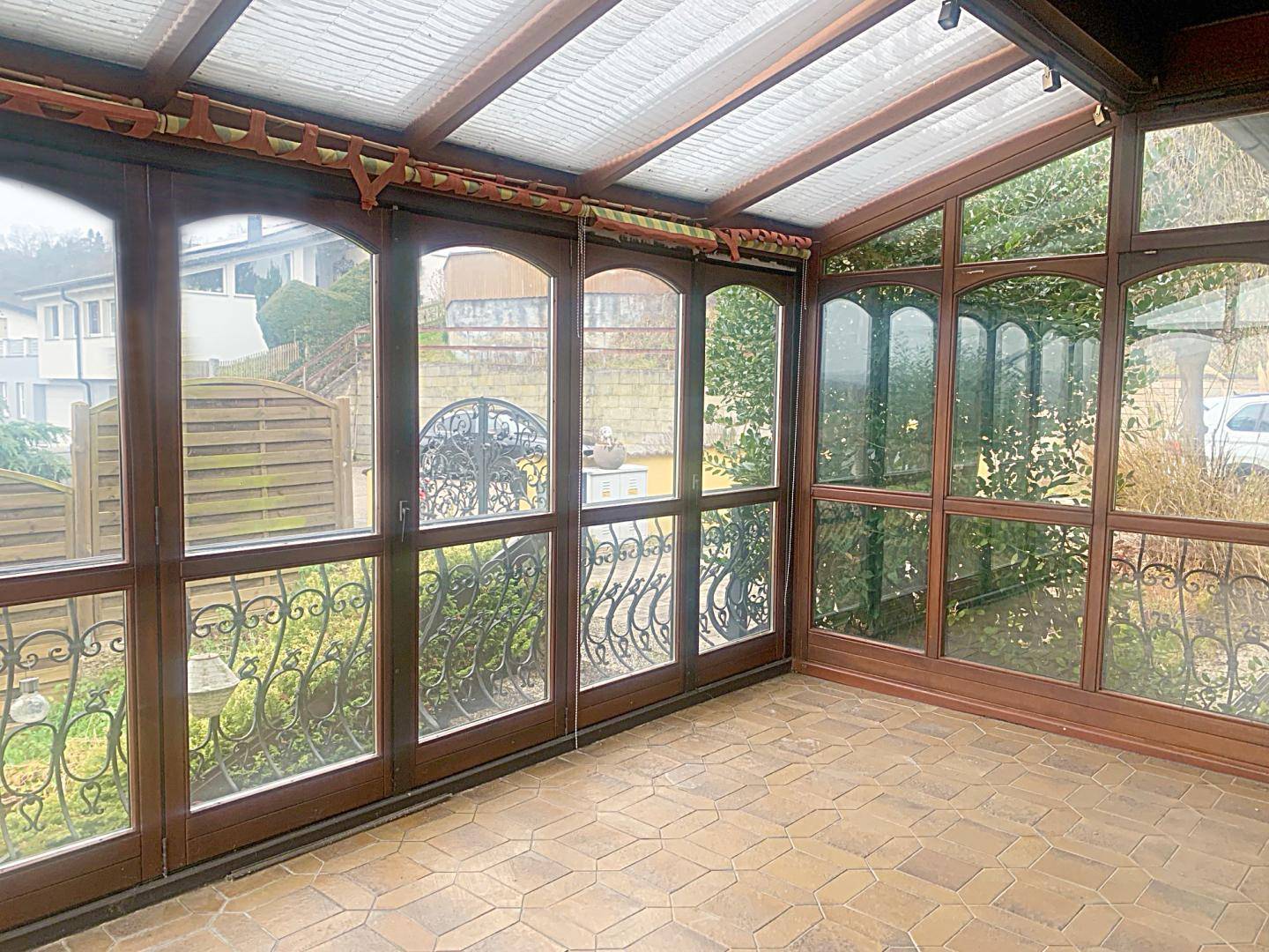 Heller Wintergarten mit großen Glasflächen, Fliesenboden und Blick in den Garten.