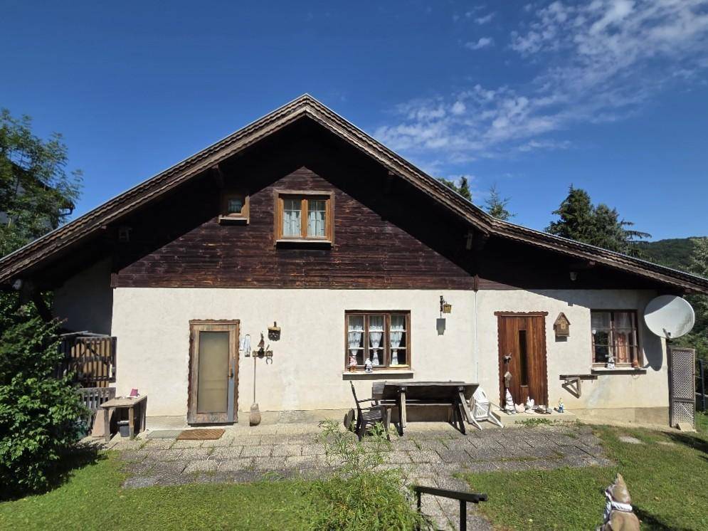 Sonniger Terrassenbereich am Haus mit Holztisch und Stühlen, umgeben von Grün.