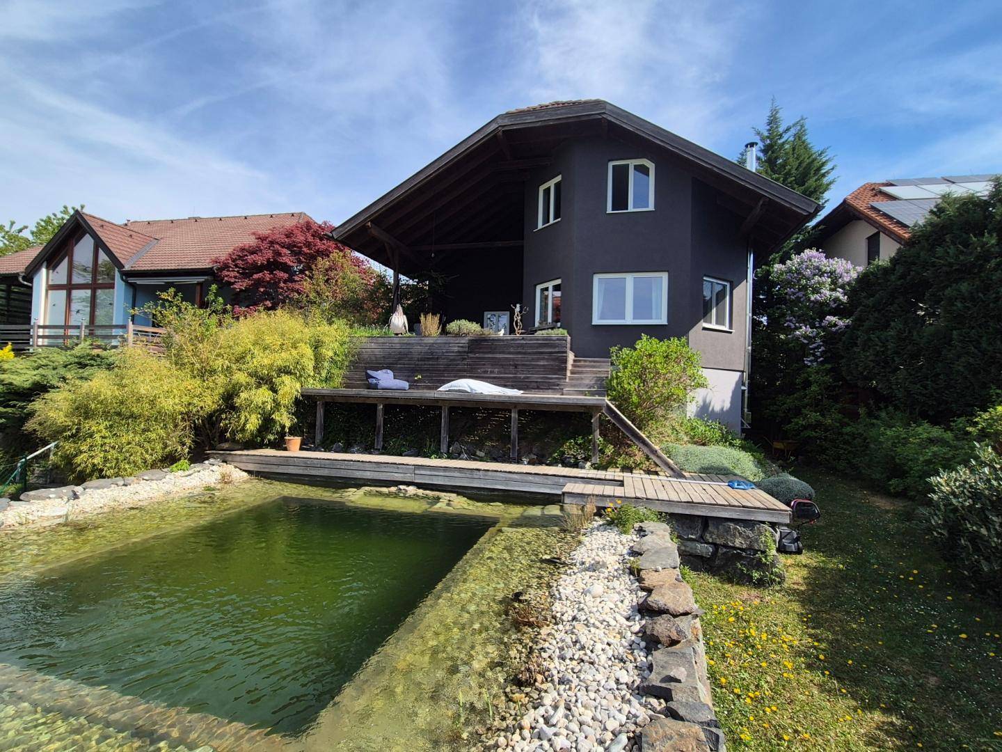 Ansicht des Hauses vom Garten aus mit Naturpool, Holzdeck und überdachtem Essbereich im Freien.