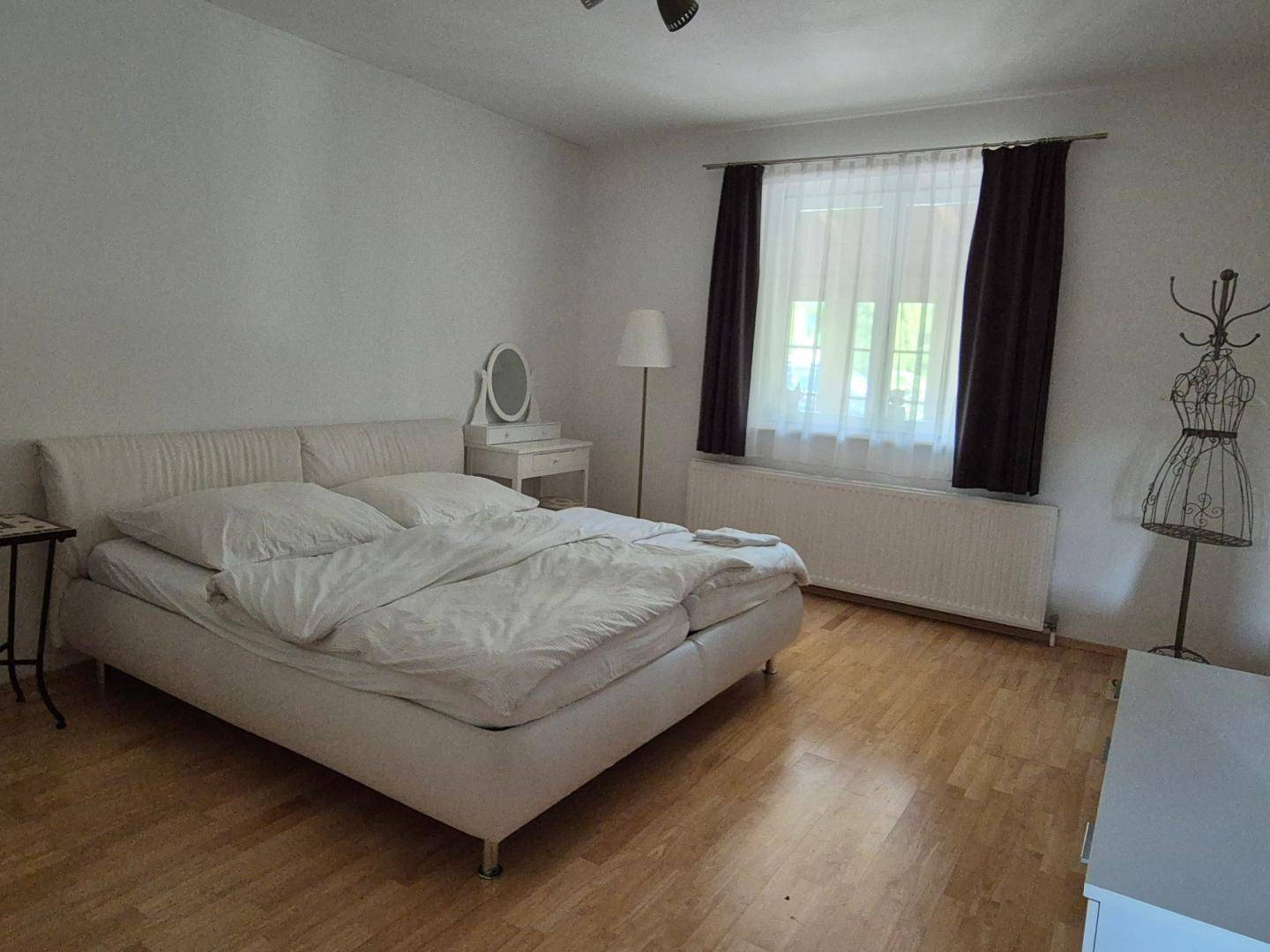 Helles Schlafzimmer mit weißem Doppelbett, Holzfußboden und einem Fenster mit Vorhängen, schlicht eingerichtet.