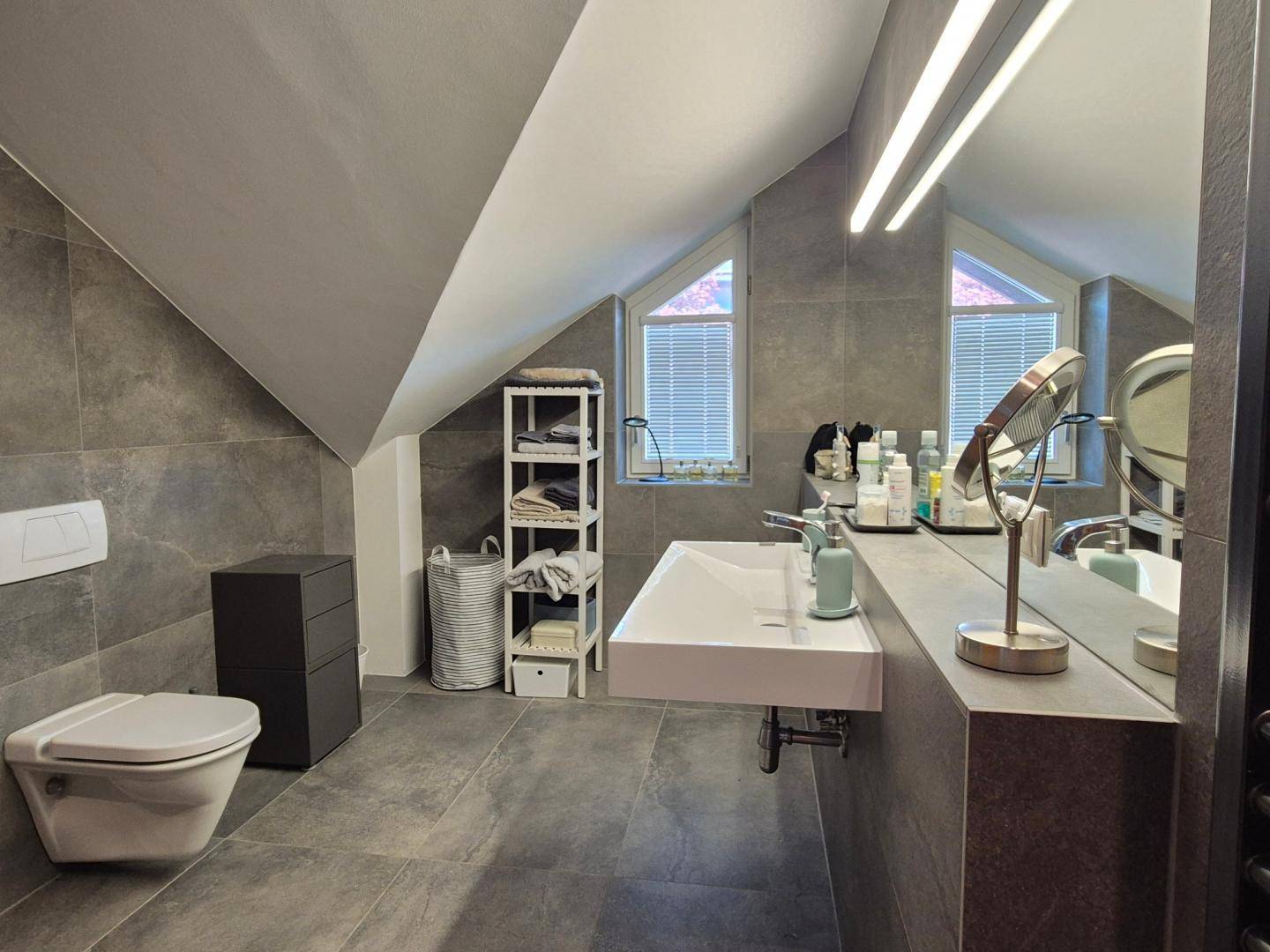 Modernes Dachgeschossbad mit grauen Fliesen, wandhängendem WC und Waschbecken unter schrägen Decken.