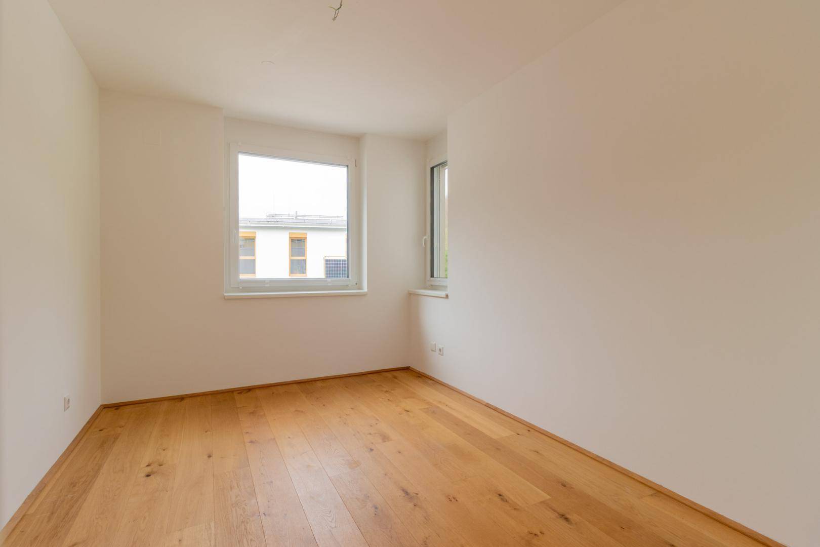 Unmöbliertes Schlafzimmer mit zwei Fenstern, Holzboden und weiß gestrichenen Wänden.