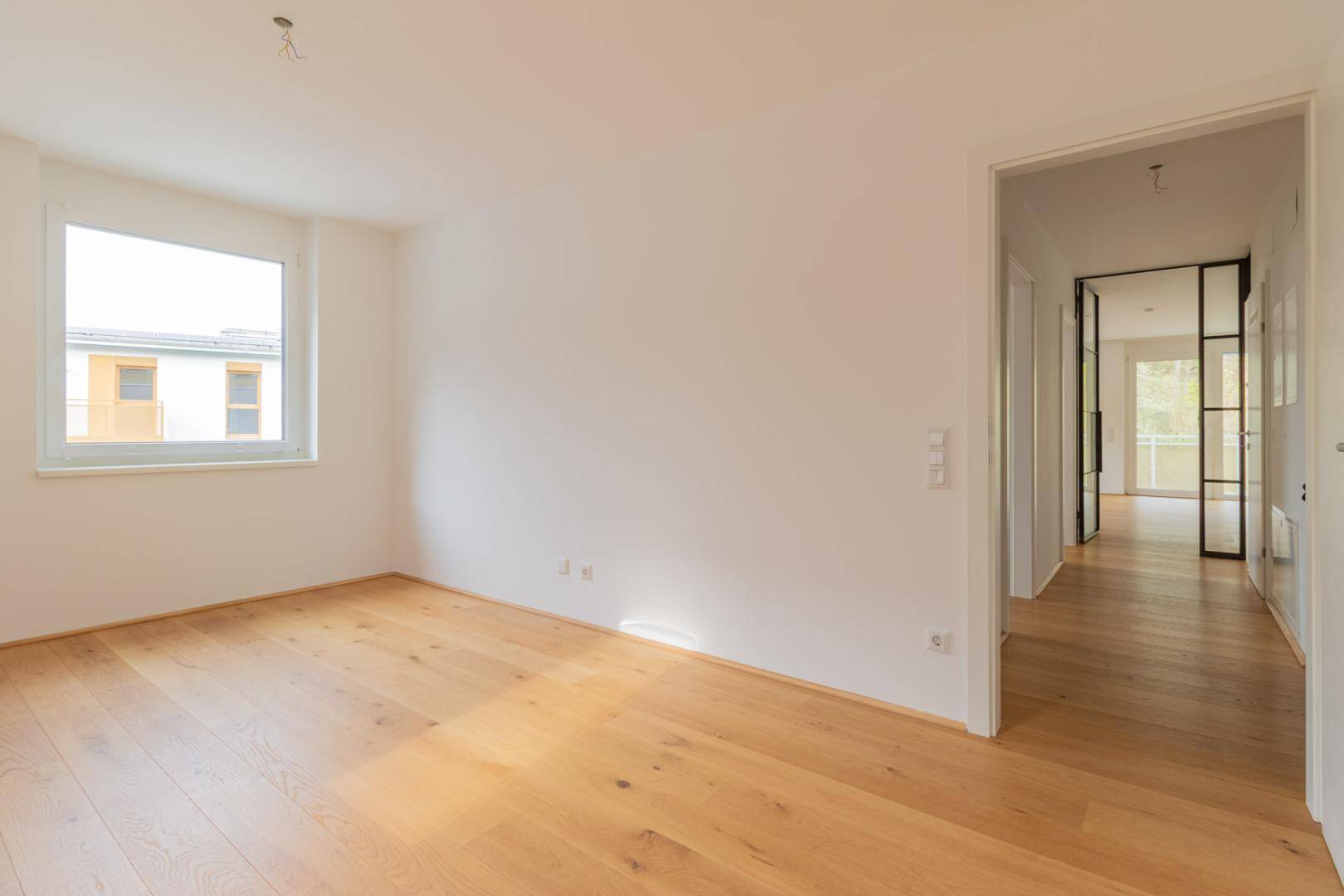 Helles, unmöbliertes Schlafzimmer mit Holzboden und Blick in den Flur mit Glastür.