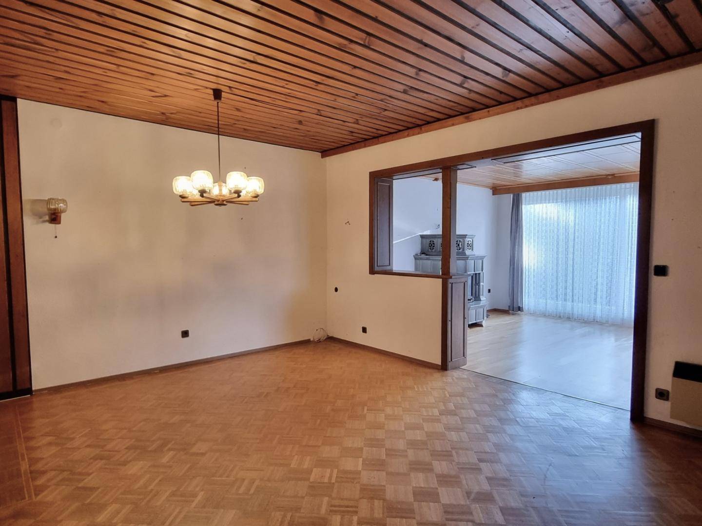 Essbereich mit Parkettboden, Holzbalkendecke und Durchgang zu einem weiteren Wohnraum mit Kachelofen.
