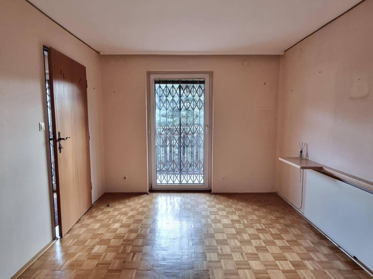 Leeres Zimmer mit Parkettboden und Zugang zu einem Außenbereich, gesichert durch ein Gitter.