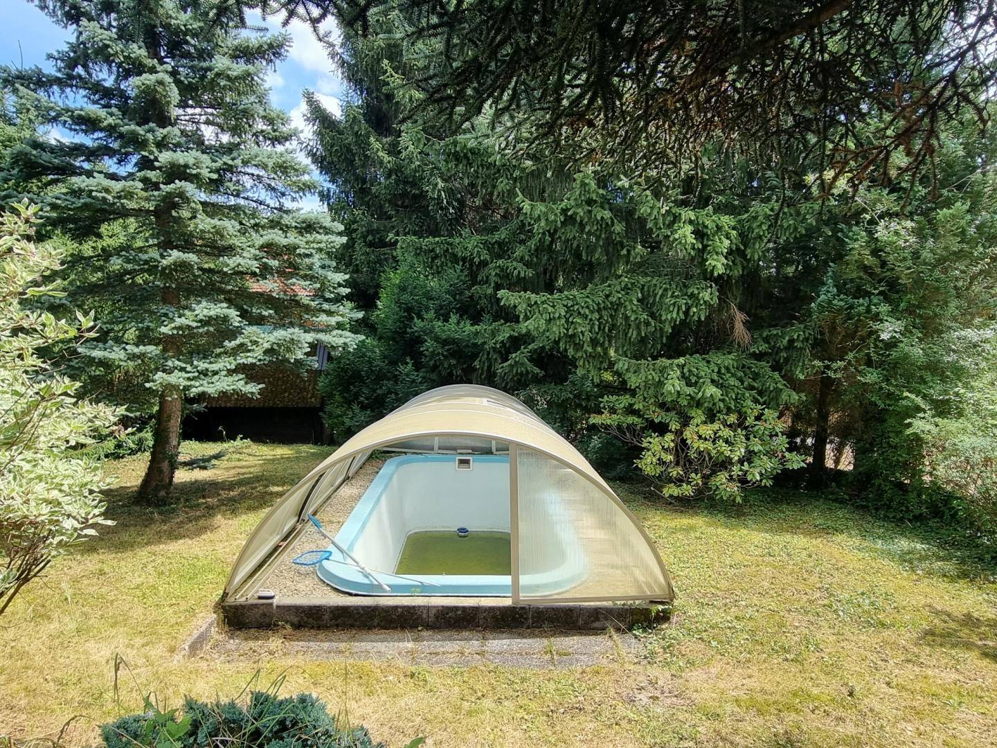 Nahaufnahme des überdachten Außenpools in einem gepflegten Garten mit Bäumen und Rasenfläche.