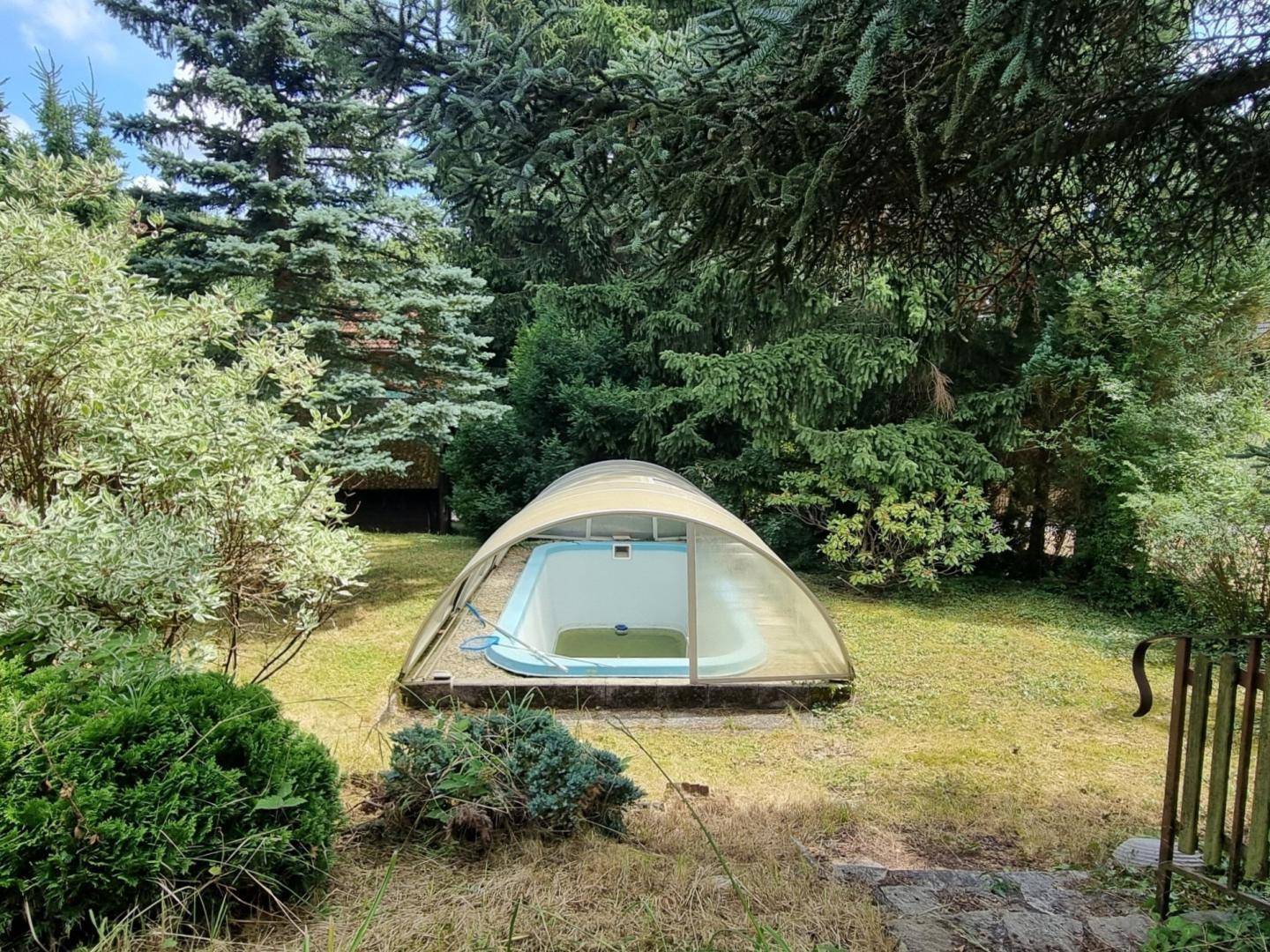 Überdachter Außenpool in einem grünen Garten, umgeben von hohen Nadelbäumen und Büschen.