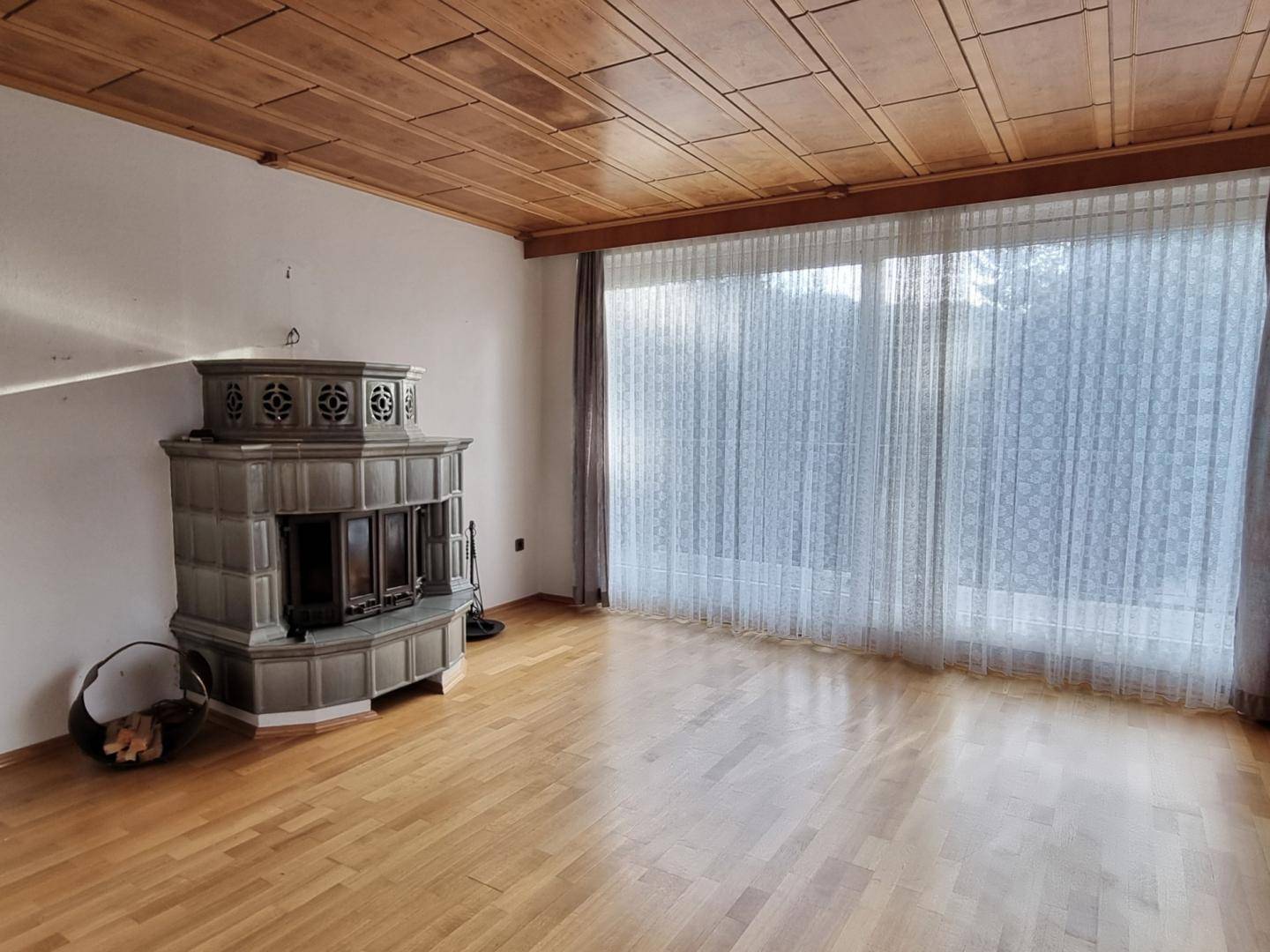 Geräumiges Wohnzimmer mit traditionellem Kachelofen, Holzboden und großen Fenstern für viel Tageslicht.