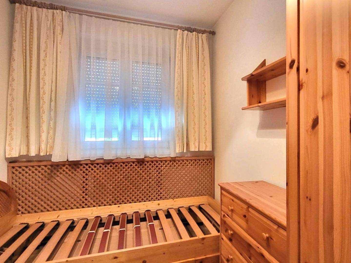 Helles Schlafzimmer mit Einzelbett, Holzmöbeln und Fenster mit Rollladen.