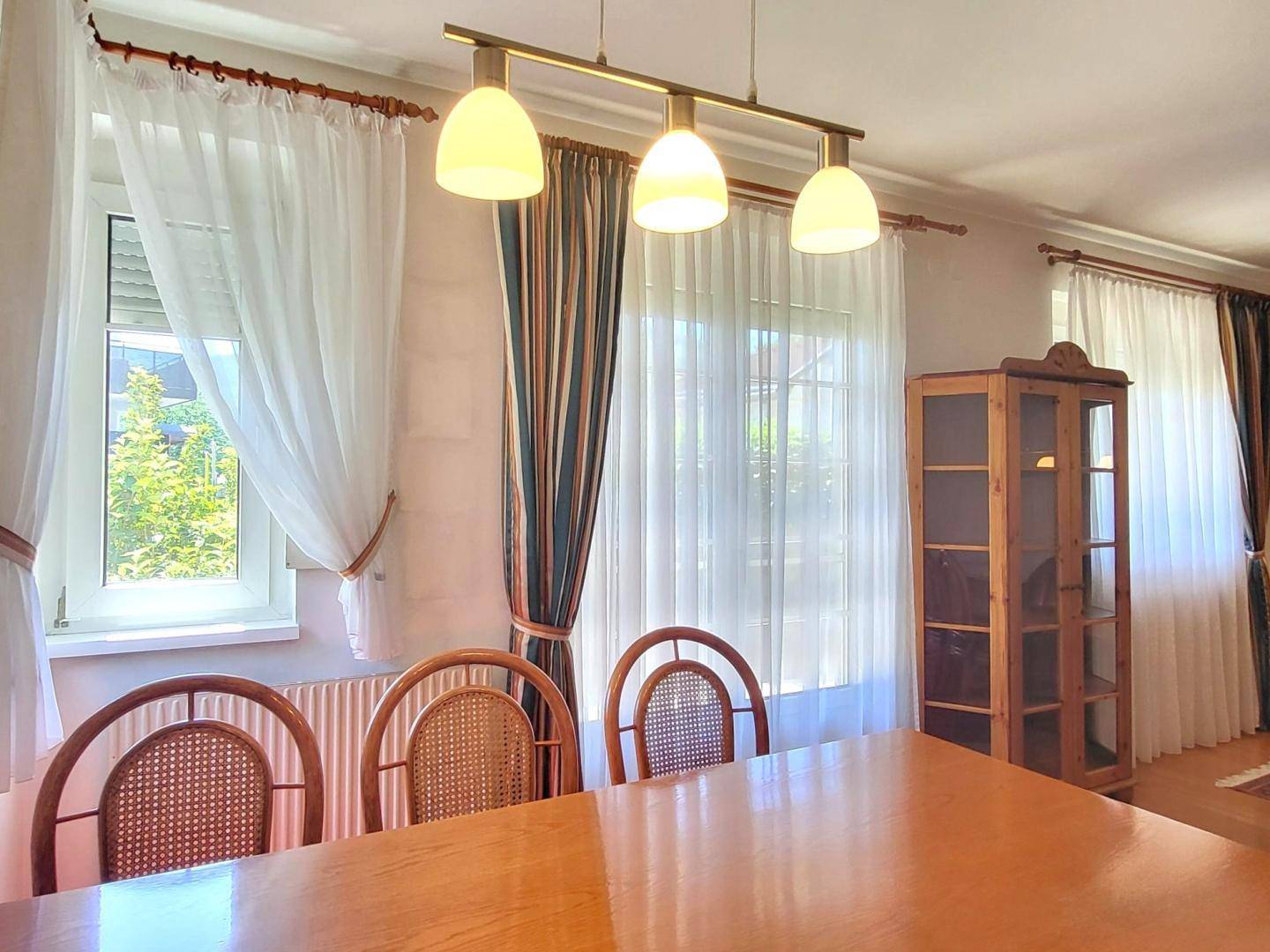 Heller Essbereich mit Holztisch, Korbstühlen und großen Fenstern mit Vorhängen.
