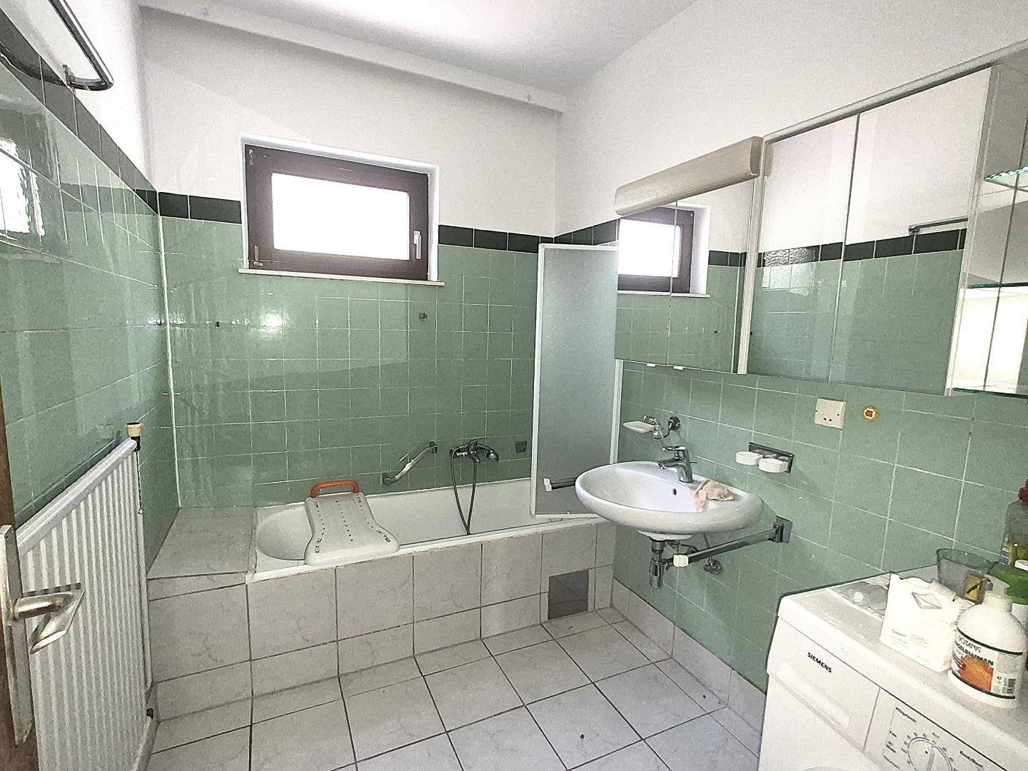 Badezimmer mit grünen Fliesen, Badewanne, Waschbecken und einem Fenster, im Stil der 80er Jahre.