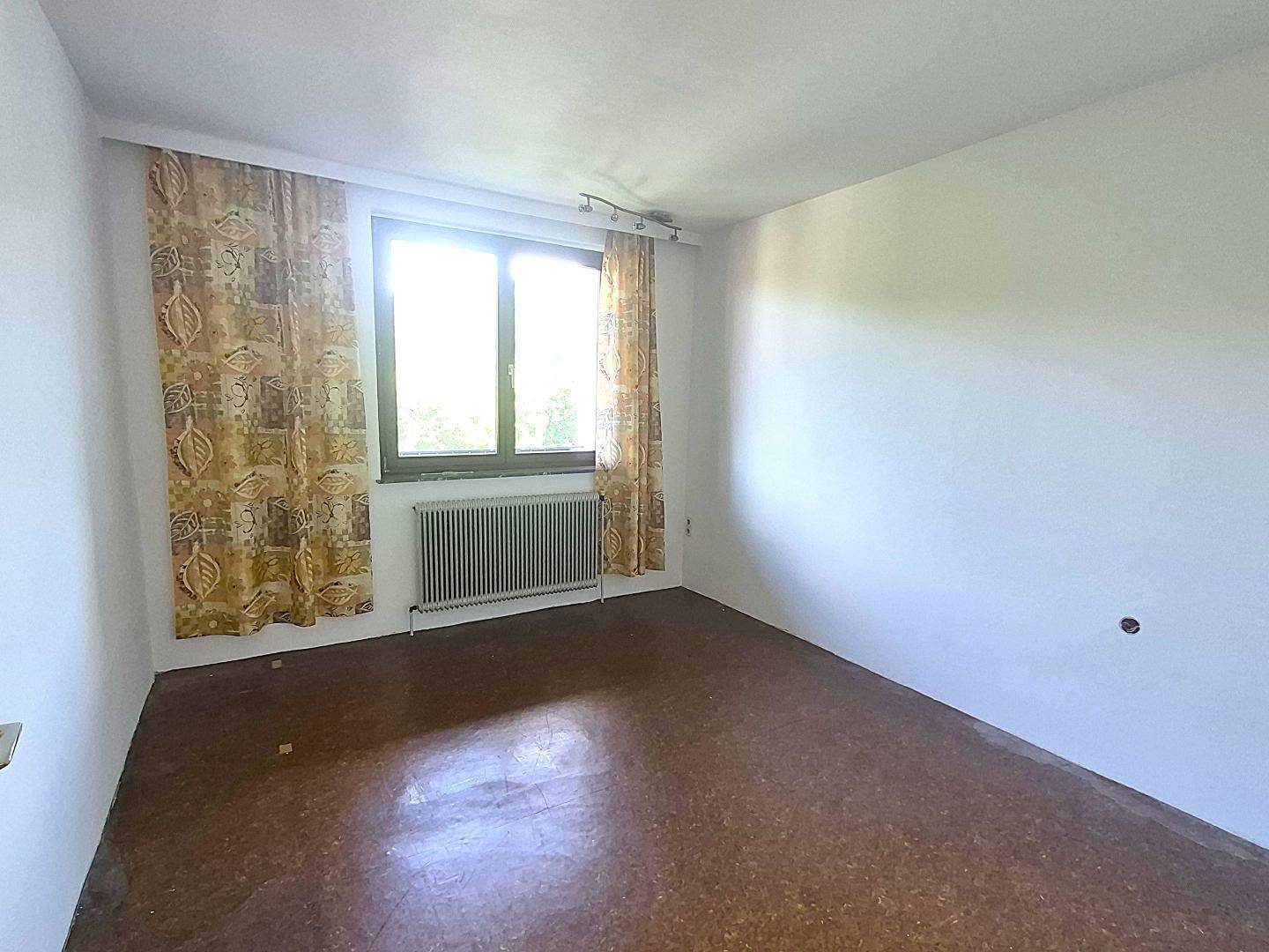 Helles Zimmer mit einem Fenster, Heizkörper und einem braunen Linoleumboden, bereit zur Renovierung.