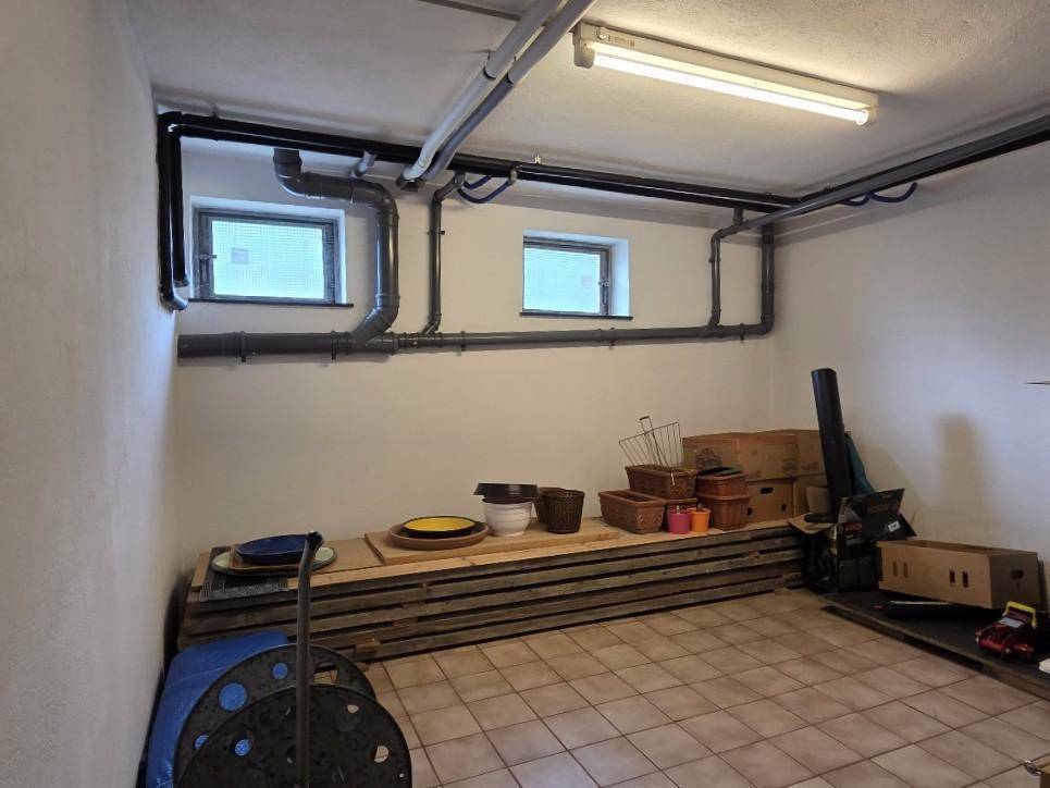 Kellerraum mit sichtbaren Rohren, kleinen Fenstern und Fliesenboden, genutzt als Abstellraum.