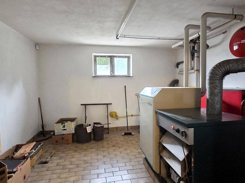 Heizungsraum im Keller mit großem Heizkessel, sichtbaren Rohren und Fliesenboden.