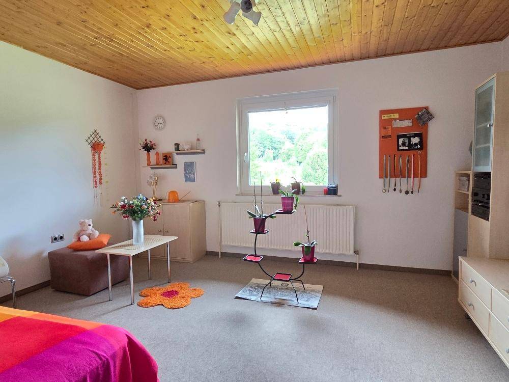 Helles Zimmer mit Holzdecke, Teppichboden und Fenster mit Blick ins Grüne.