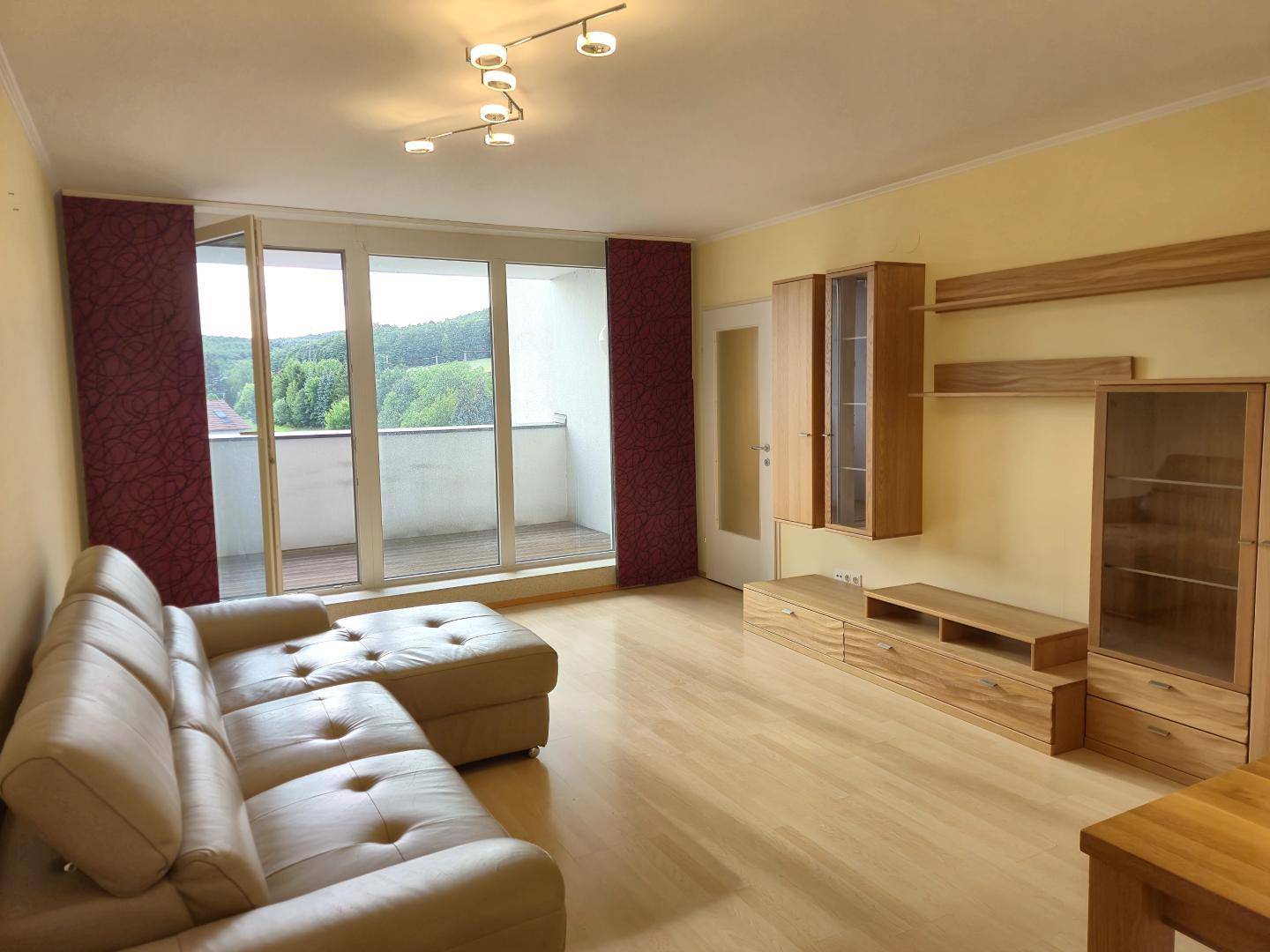 Helles Wohnzimmer mit bequemer Couch, Holzmöbeln und direktem Zugang zum Balkon mit Grünblick.
