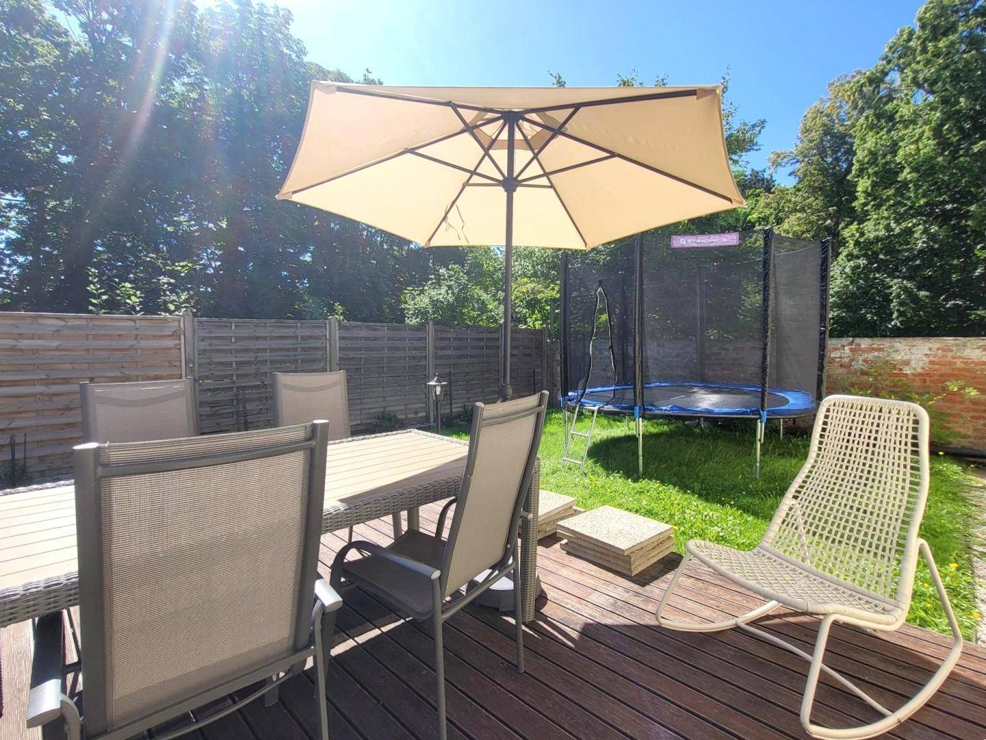 Sonniger Terrassenbereich mit Esstisch, Sonnenschirm und einem Trampolin im Garten.