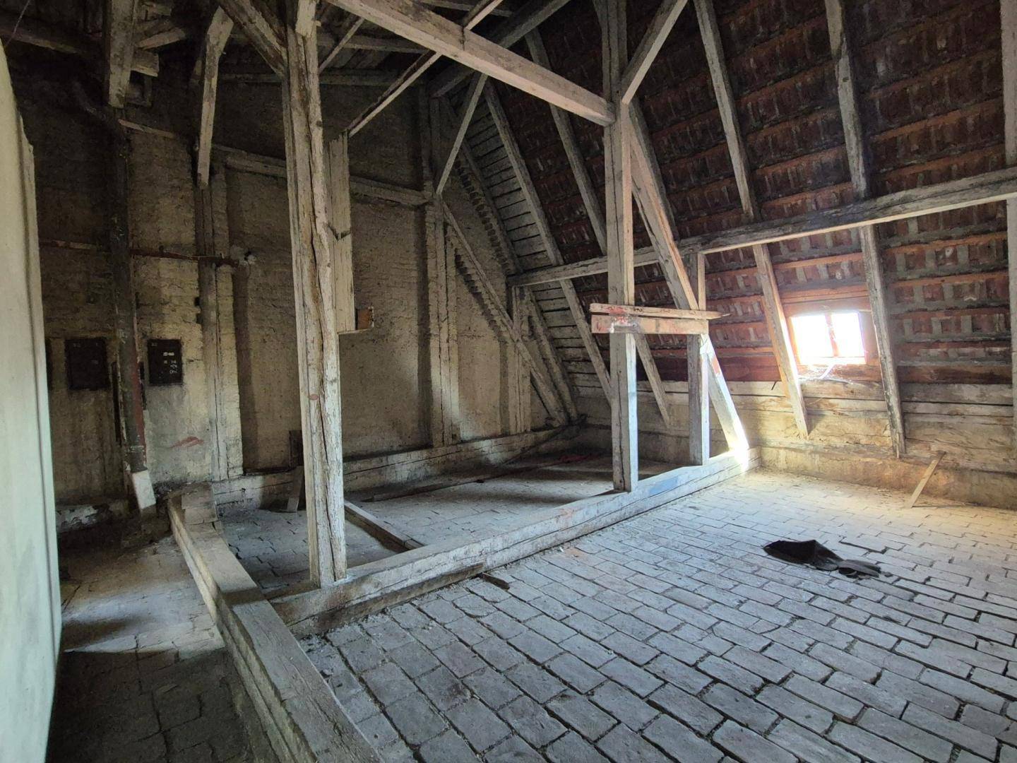 Unausgebauter Dachboden mit sichtbaren Holzbalken, Ziegelboden und kleinem Fenster.