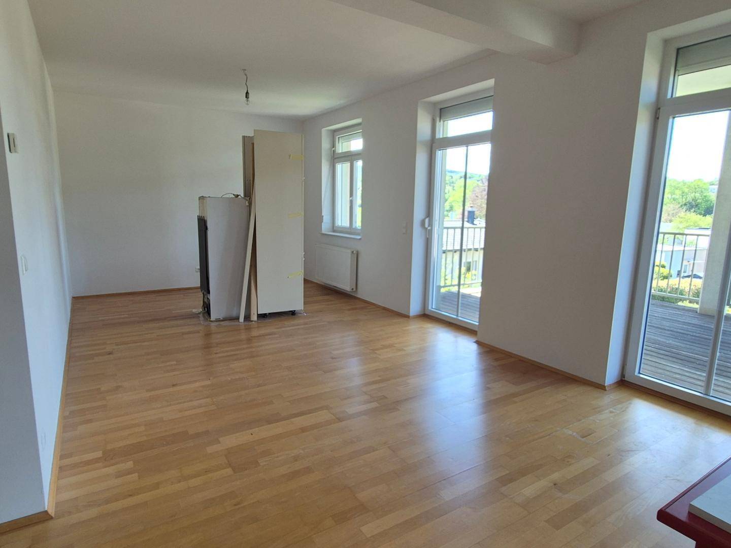 Heller Wohnraum mit Holzboden und großen Fenstern, die Zugang zu einem Balkon bieten.