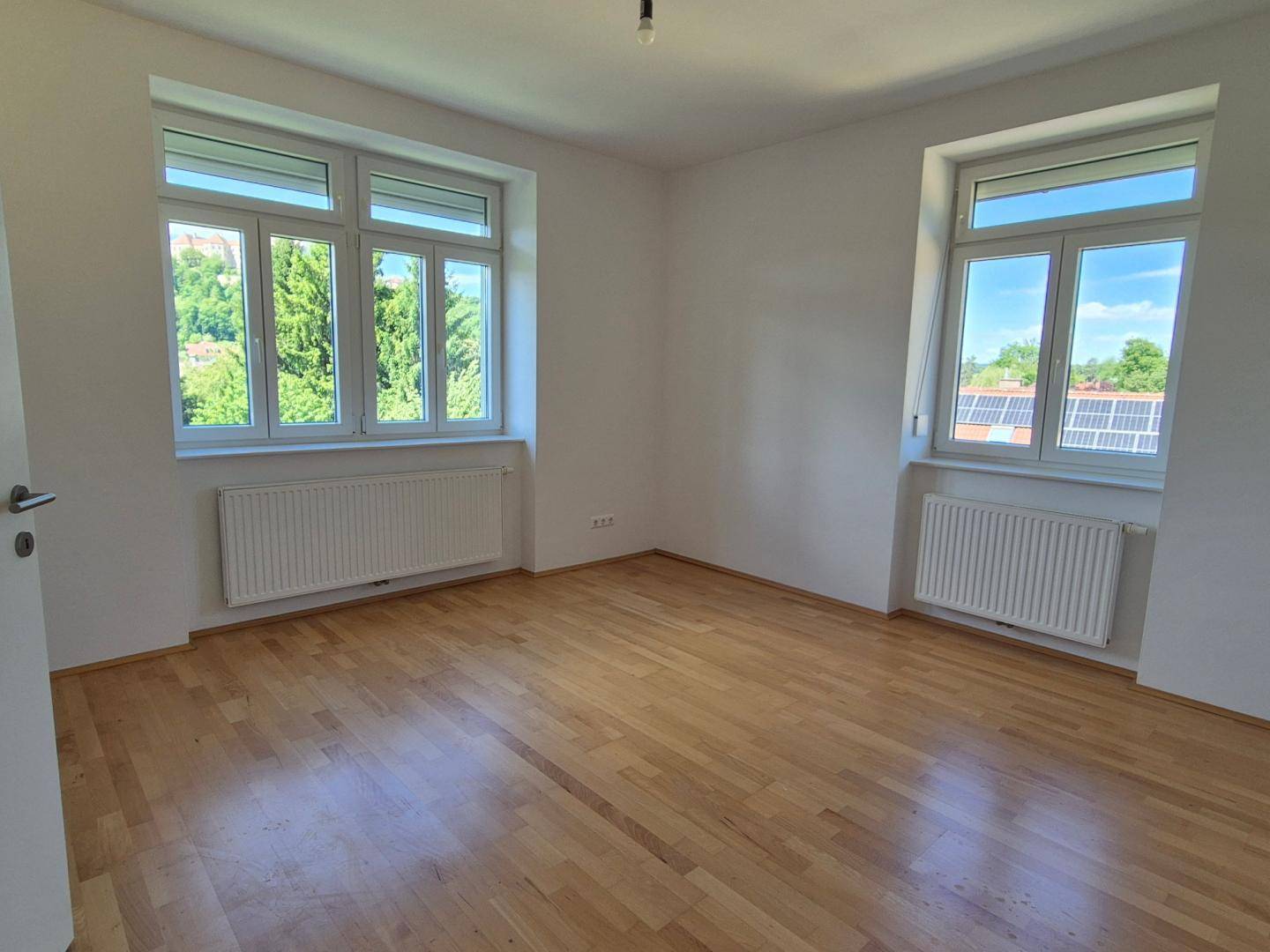 Geräumiges Zimmer mit Holzboden, weißen Wänden und Fenstern mit grüner Aussicht.