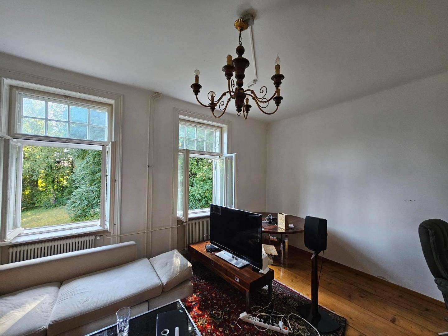 Wohnzimmer mit Holzdielenboden, Sofa und einem traditionellen Kronleuchter, hell durch Fenster.