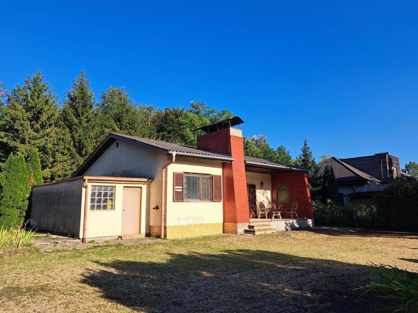 Gesamtaufnahme des Hauses mit Garten, Anbau und umliegenden Bäumen unter blauem Himmel.