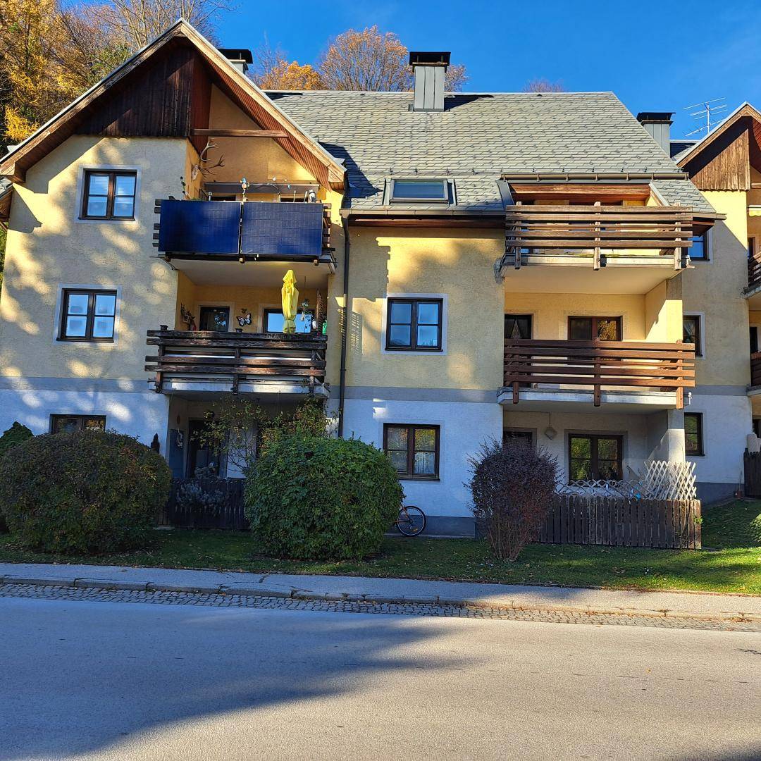 Mehrfamilienhaus mit gelber Fassade, Holzbalkonen und Satteldach in einer grünen Umgebung.
