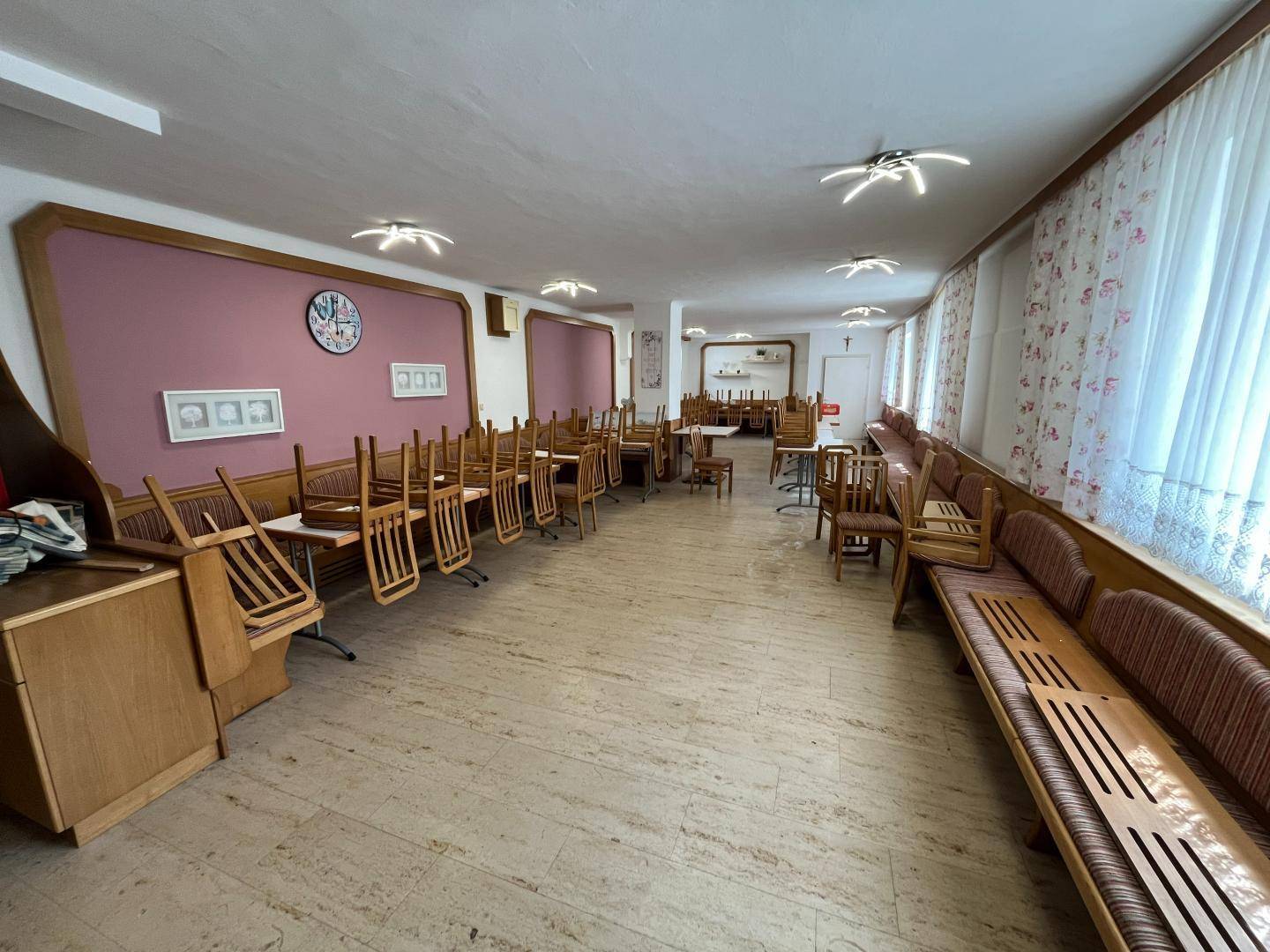Großer, leerer Speisesaal mit gestapelten Stühlen, gepolsterten Bänken und hellen Vorhängen.