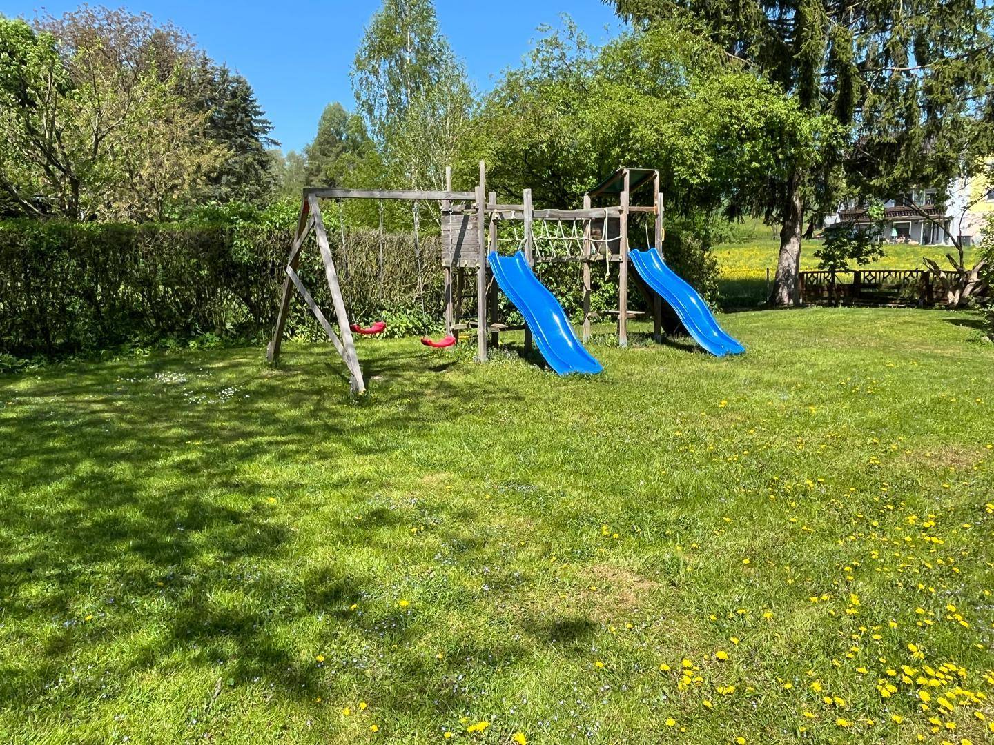Grüner Gartenbereich mit einem Holzspielplatz, Rutschen und Schaukeln unter blauem Himmel.
