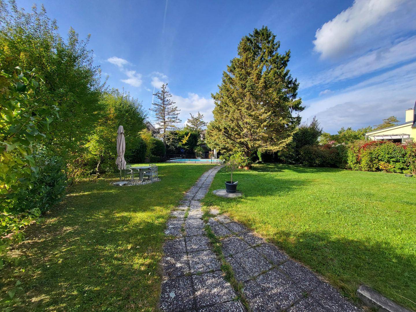 Weitläufiger Garten mit grünem Rasen, altem Baumbestand und einem gepflasterten Weg zum Pool.