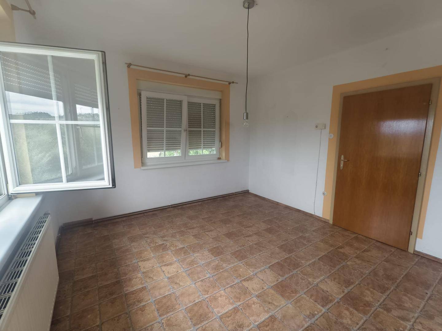 Zimmer mit braunen Fliesen, weißen Wänden und zwei Fenstern mit Jalousien, hell und funktional.