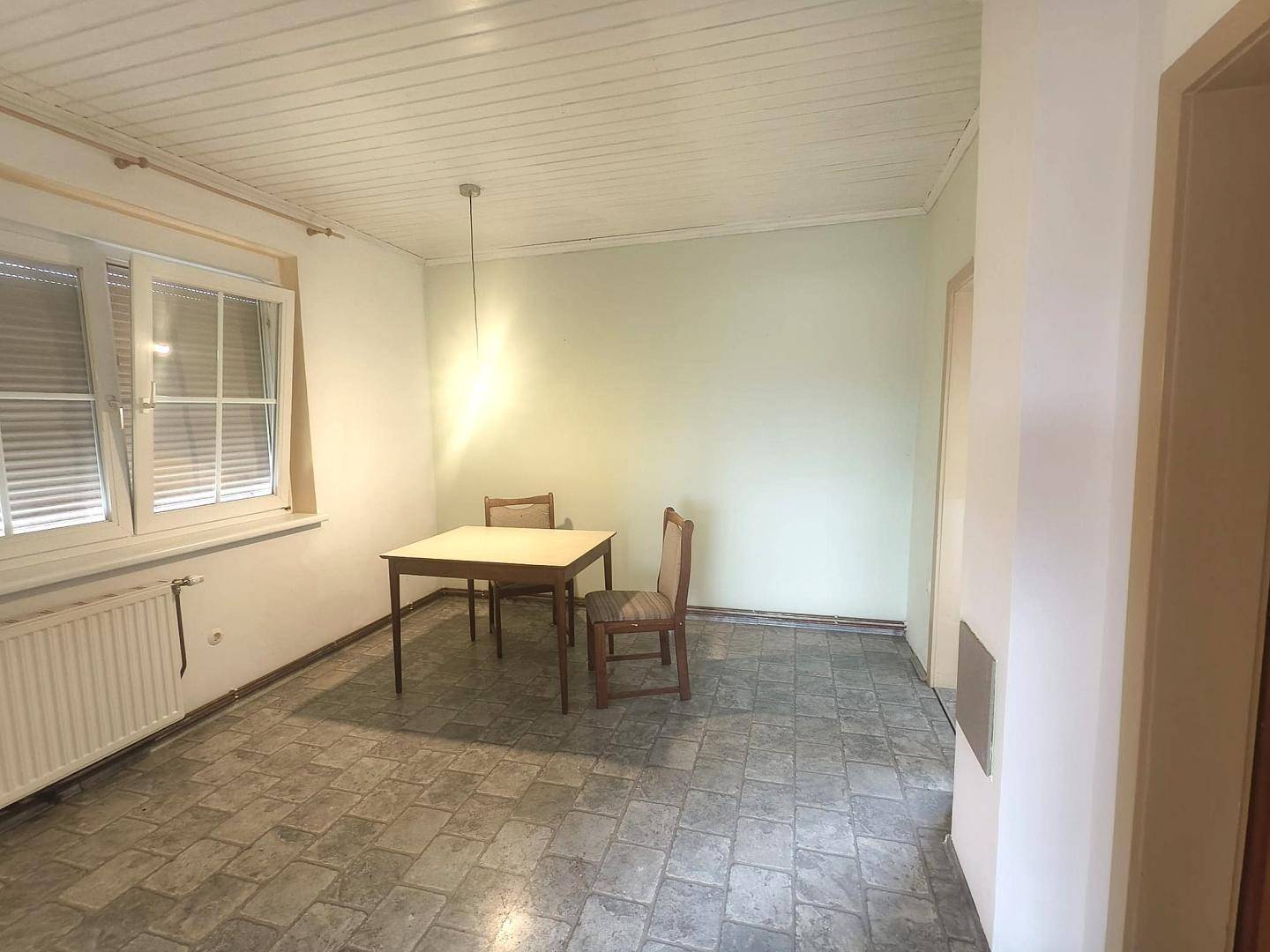 Esszimmer mit kleinem Holztisch, zwei Stühlen und Fenstern mit Jalousien, auf Fliesenboden.