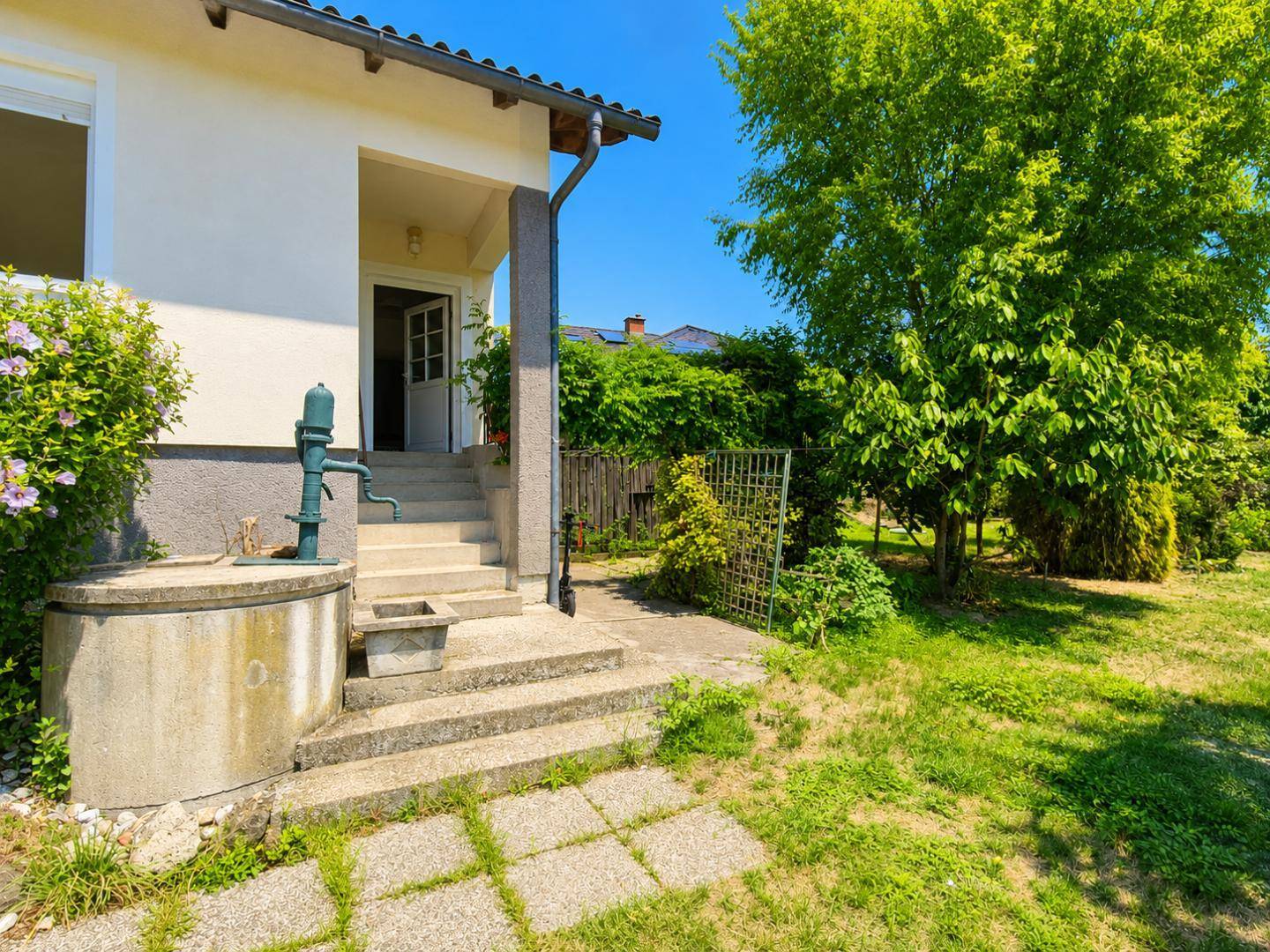 Eingangsbereich des Hauses mit Steintreppe, alter Wasserpumpe und gepflegtem Rasen im Garten.