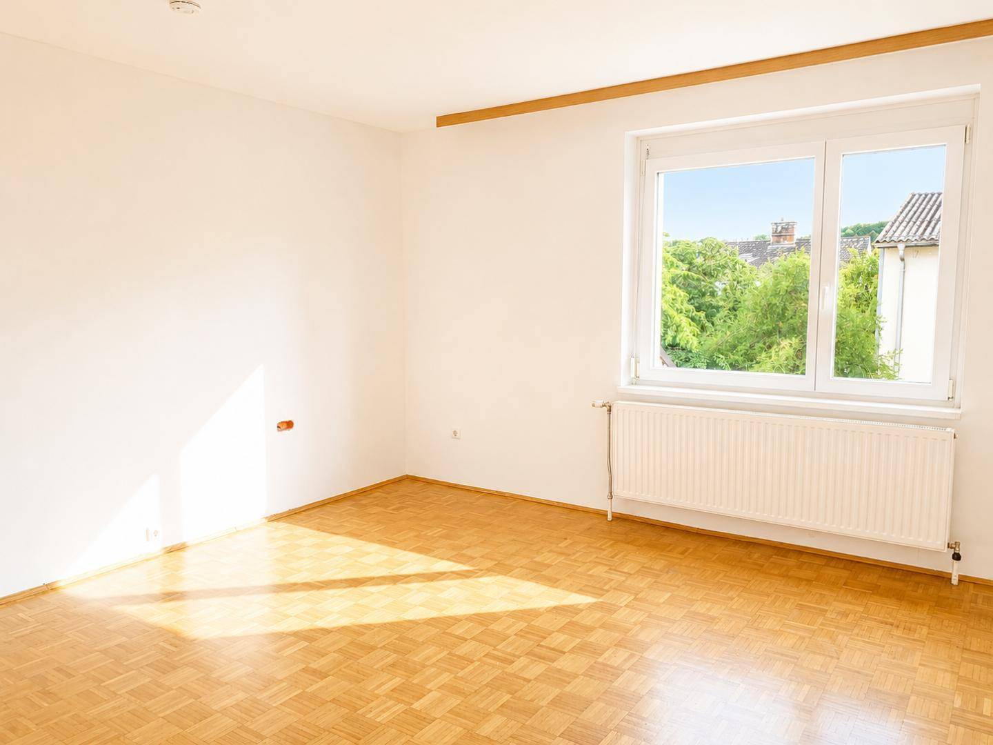 Geräumiges Zimmer mit Parkettboden, großem Fenster und Blick ins Grüne.