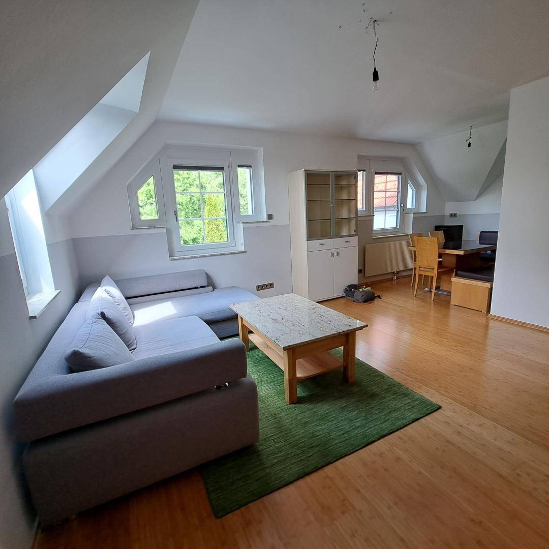 Gemütlicher Wohnbereich mit L-förmigem Sofa, Holzboden und einem Essbereich im Hintergrund unter Dachschrägen.