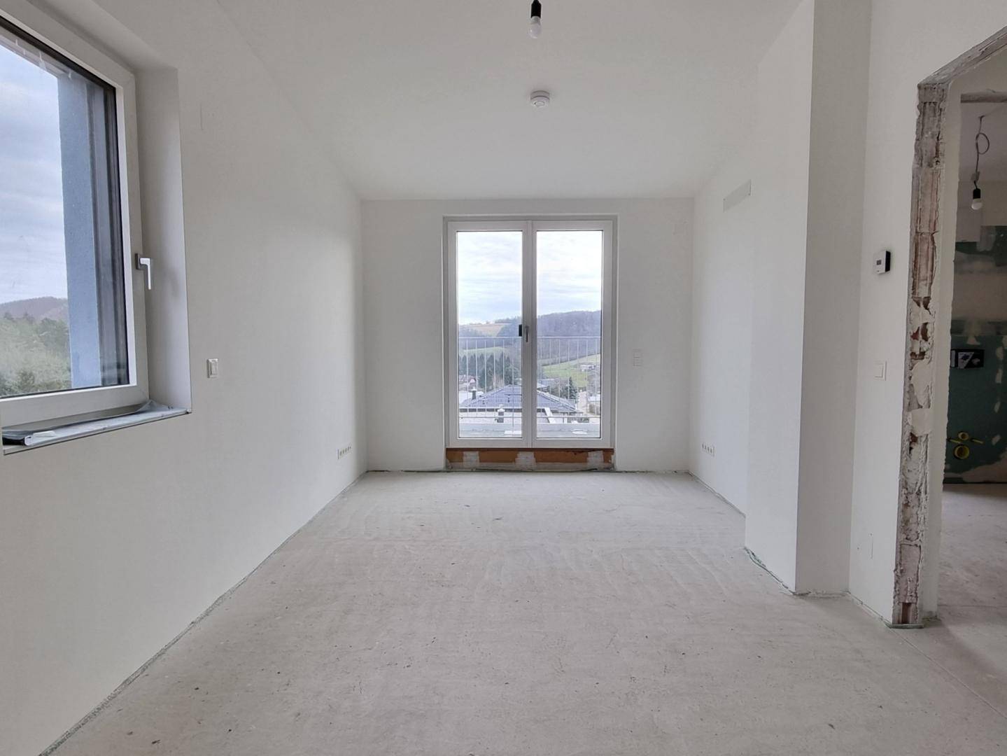 Unmöblierter Raum im Rohbau mit Balkonzugang und Blick auf die umliegende Landschaft.