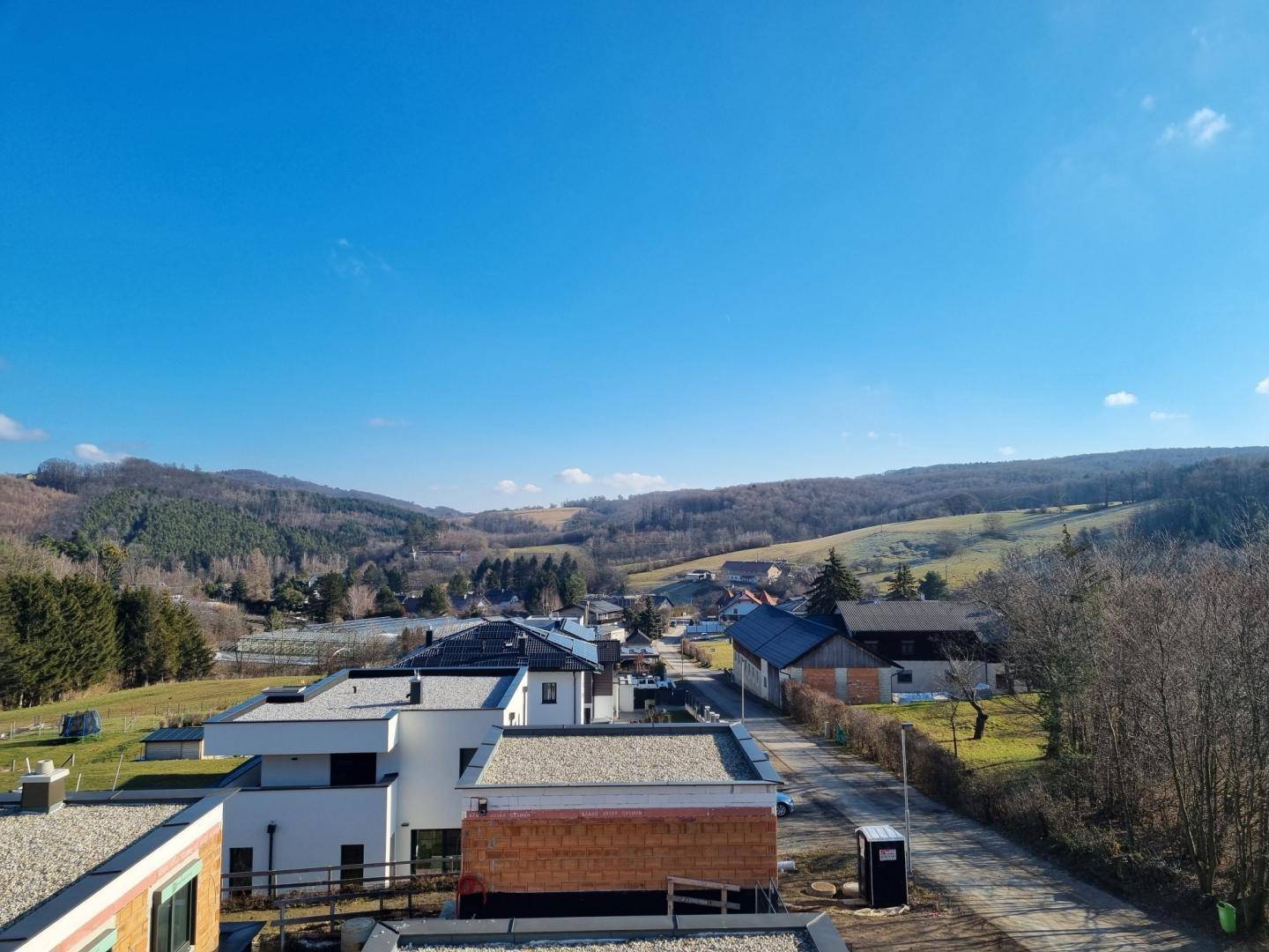 Weitläufiger Blick über eine ländliche Siedlung mit Häusern und grünen Hügeln unter blauem Himmel.