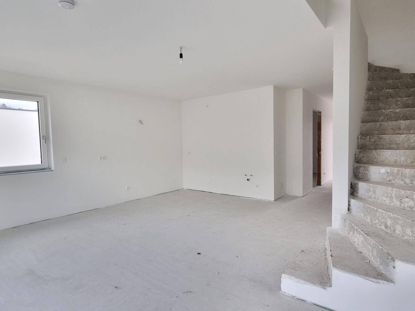 Offener Wohnbereich im Rohbau mit Treppenaufgang und einem Fenster, das Tageslicht spendet.