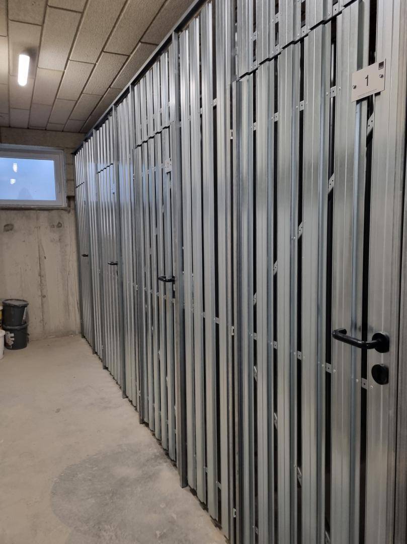 Reihe von abschließbaren Lagerabteilen aus Metall in einem Kellerraum mit kleinem Fenster.