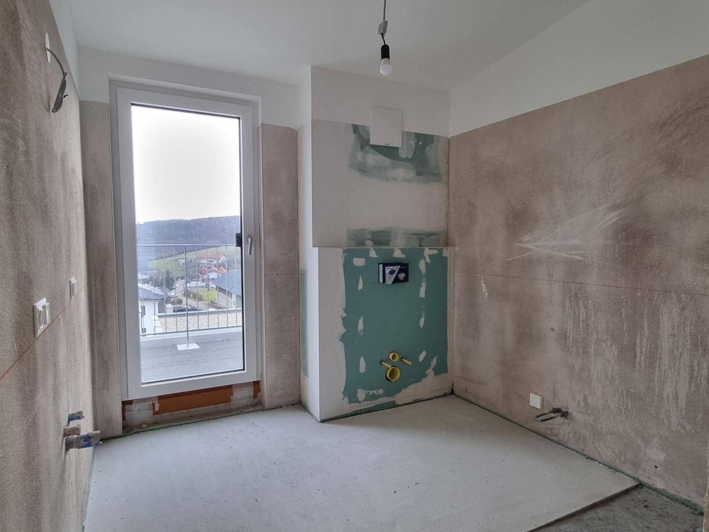 Unfertiges Badezimmer im Rohbau mit vorbereiteten Anschlüssen und einem Fenster.