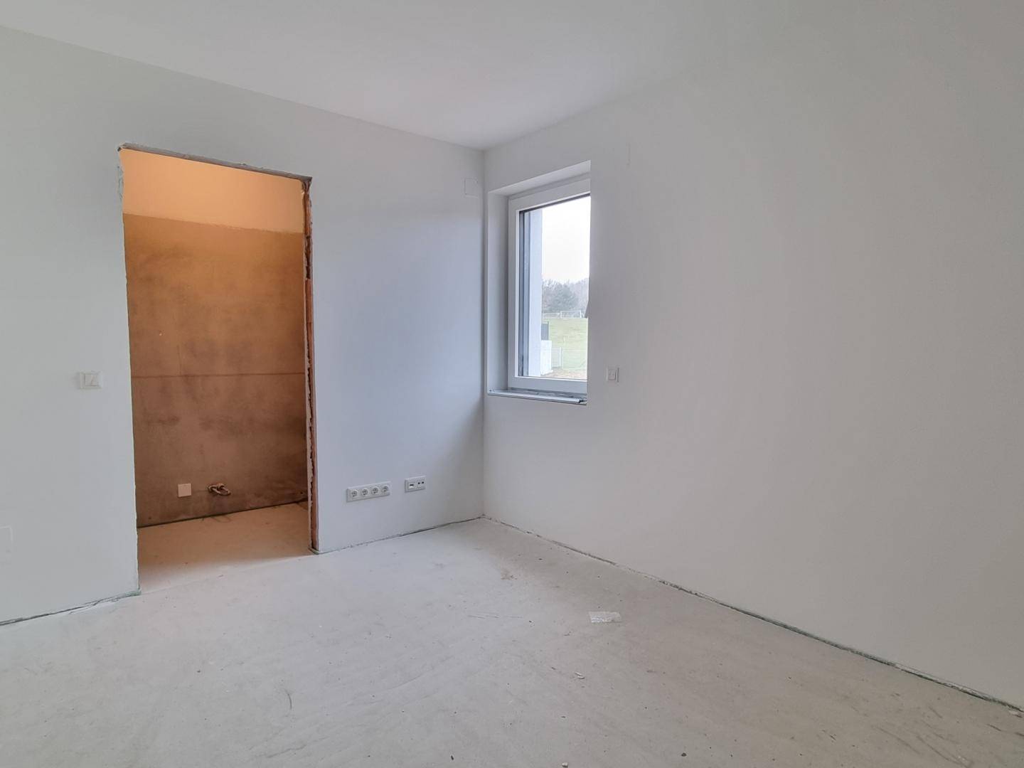 Unmöblierter Raum im Rohbau mit einem Fenster und einer Öffnung zu einem weiteren Bereich.