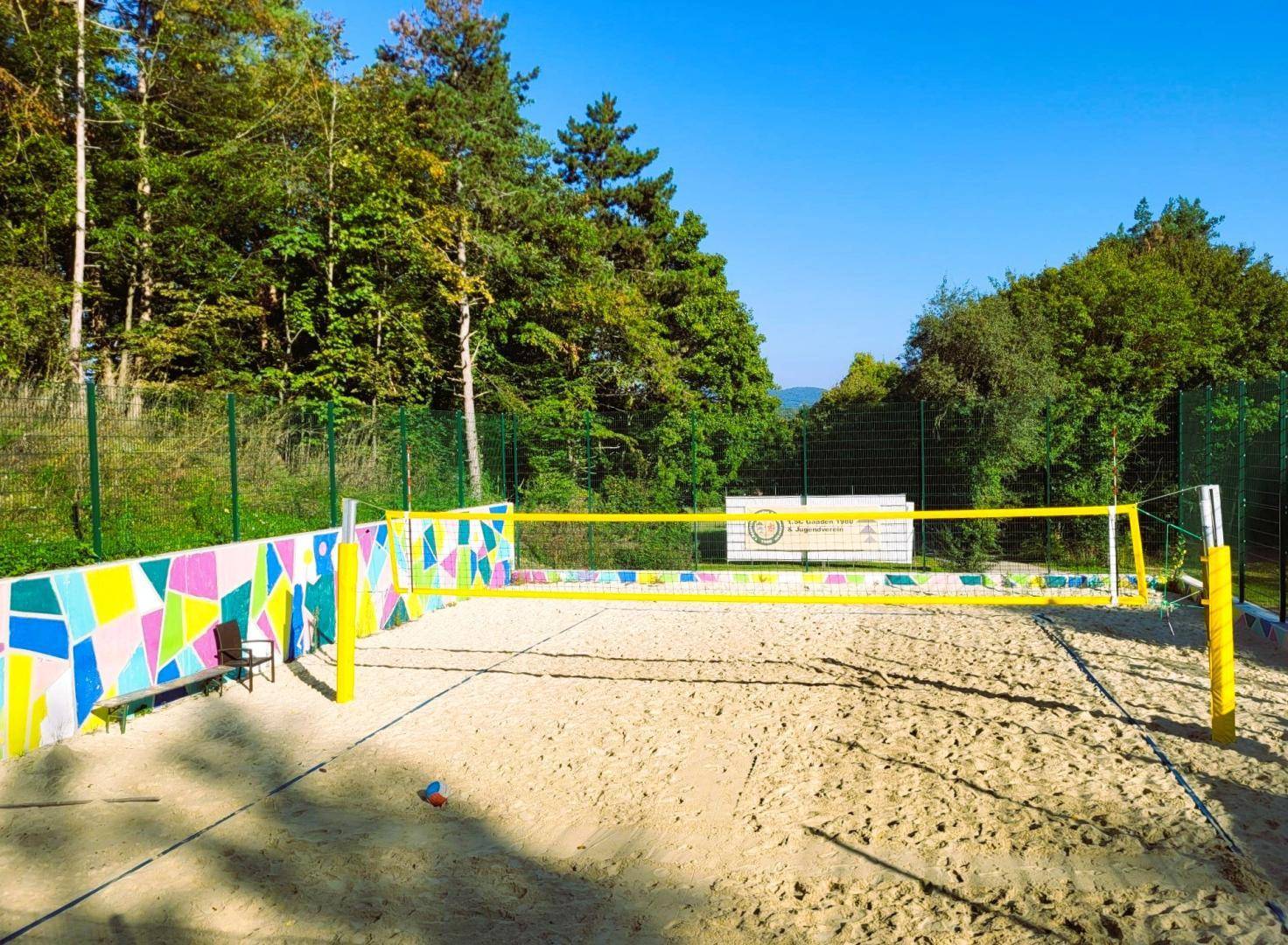 Sandvolleyballfeld mit bunter Mosaikwand und umgebenden Bäumen unter klarem Himmel.