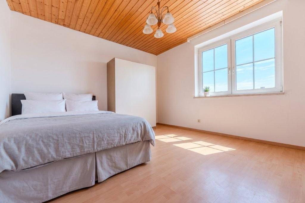 Helles Schlafzimmer mit Holzbalkendecke, hellem Laminatboden und einem großen Fenster, das viel Tageslicht hereinlässt.