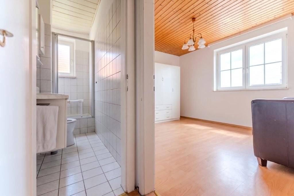 Blick in ein Badezimmer mit weißen Fliesen, Toilette und Dusche, angrenzend an einen Raum mit Holzdecke.